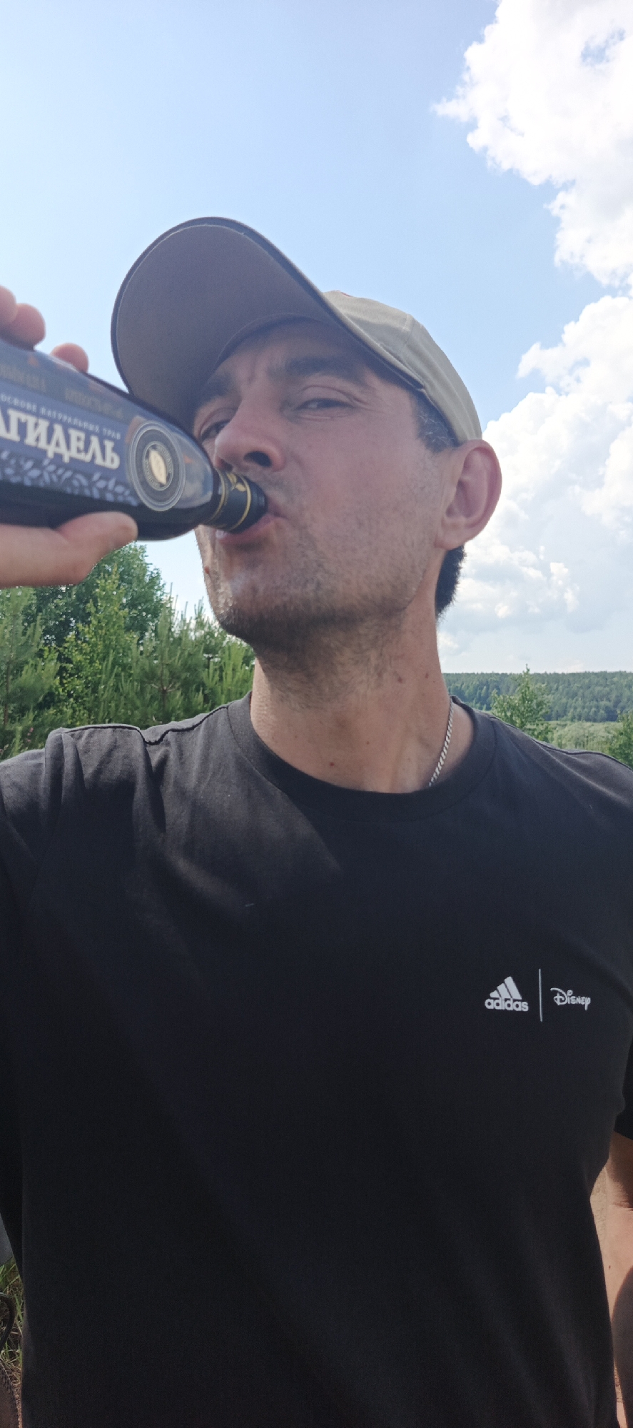 A close-up, low-angle outdoor selfie shows a man wearing a black Adidas x Disney t-shirt and a tan baseball cap. He is holding a dark bottle with a label in Russian and drinking from it. His mouth is slightly open as he sips from the bottle. The background consists of a bright blue sky with scattered white clouds, green trees, and a distant hillside covered in trees. The image appears to have been taken during the day.