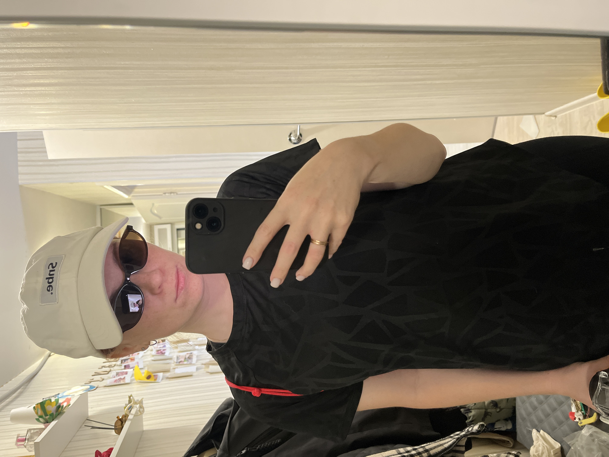 A young woman wearing a white baseball cap, sunglasses, and a black t-shirt is taking a mirror selfie. She is standing in what appears to be a room with a closet and shelves in the background. The wall behind her has vertical stripes. The woman is holding a black smartphone in her right hand. A red strap is visible over her left shoulder, suggesting she might be wearing a backpack or bag. The image is taken indoors, likely in a residential setting. There is no indication of the specific location within Chaikovsky, Russia.