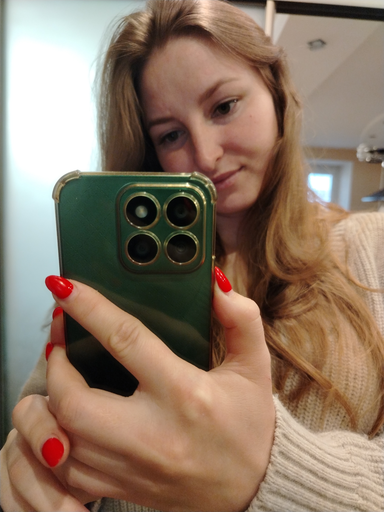 The image is a close-up selfie taken from a low angle, focusing on a young woman's face and hands holding a smartphone. The woman has long, light brown hair that falls over her shoulders and a beige knitted sweater. Her fingernails are painted a vibrant red. The smartphone she is holding is green with a gold trim around the camera module, which features four lenses arranged in a square pattern. Her face is partially obscured by the phone, but her right eye and part of her nose and mouth are visible. The background is blurred but appears to be an indoor setting with some lighting. There is no information in the image to suggest a specific location in Chaikovsky, Russia.