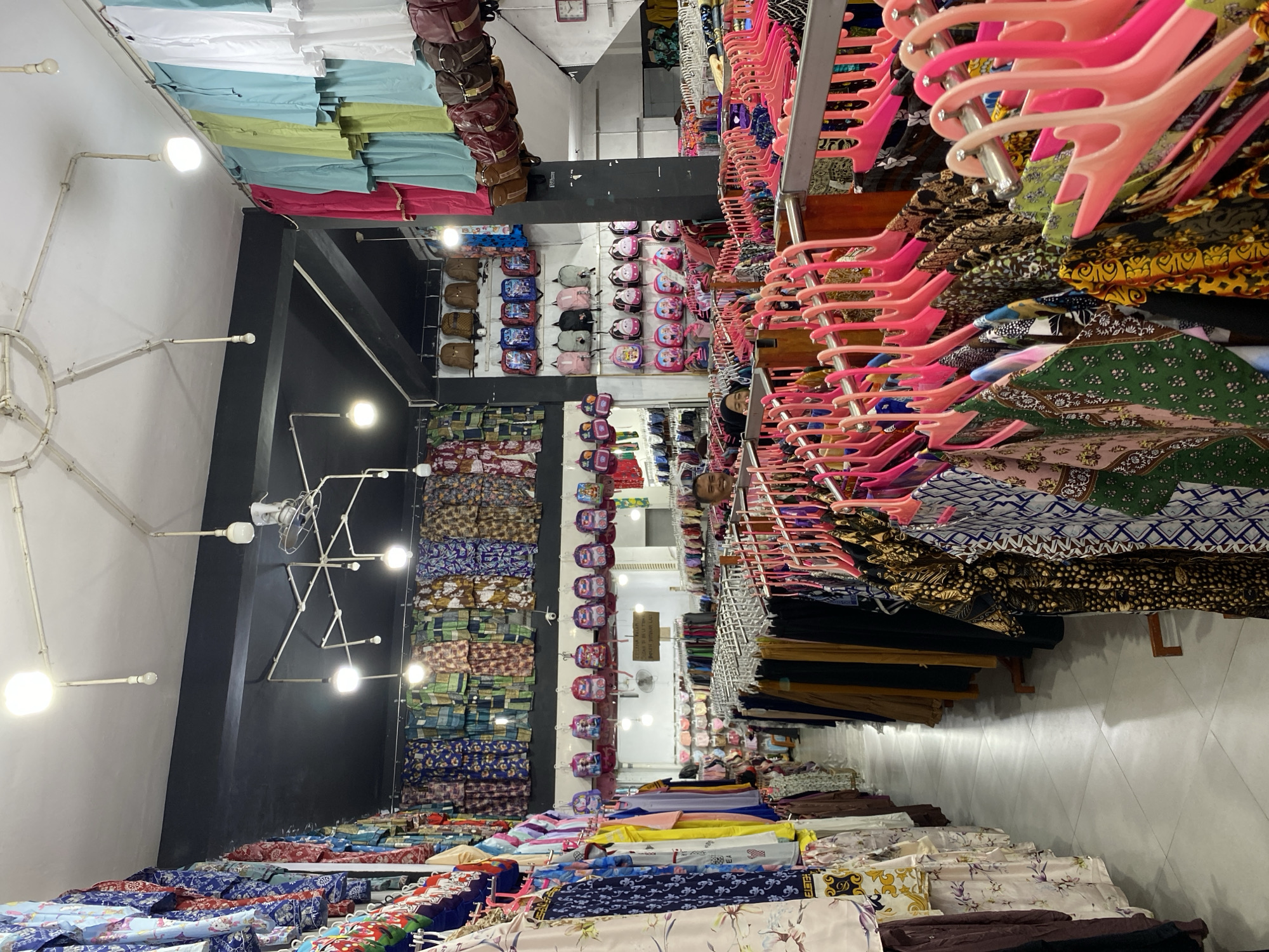 This media file depicts the interior of a retail store, likely a fabric shop or a boutique selling clothing and accessories, located in Lamteumen Barat, Indonesia. The scene is brightly lit, suggesting it is daytime, and the store appears well-stocked and organized.
The primary subjects are rows of various fabrics, garments, and accessories displayed on shelves, racks, and tables. Prominently featured are stacks of colorful fabrics with intricate patterns, as well as many pink plastic hangers holding clothing items. Several rows of backpacks, in various colors and designs, are neatly arranged on shelves, indicating a section for children's items or fashion accessories. Some handbags or purses are also visible hanging from racks.
The overall activity is the display and sale of merchandise. There are no people directly visible engaging in the shopping activity, but a few reflections in mirrors or glass surfaces might hint at the presence of staff or customers not clearly in frame. The setting is a commercial space with lighting fixtures and shelving units.
Notable details include the sheer volume and variety of textiles, suggesting a wide selection for customers. The use of colorful displays and organized shelving creates a vibrant and inviting atmosphere for shoppers. The presence of Indonesian text on a sign in the background, though illegible in this resolution, confirms the location context. The image captures a typical retail environment focused on apparel and fabrics.