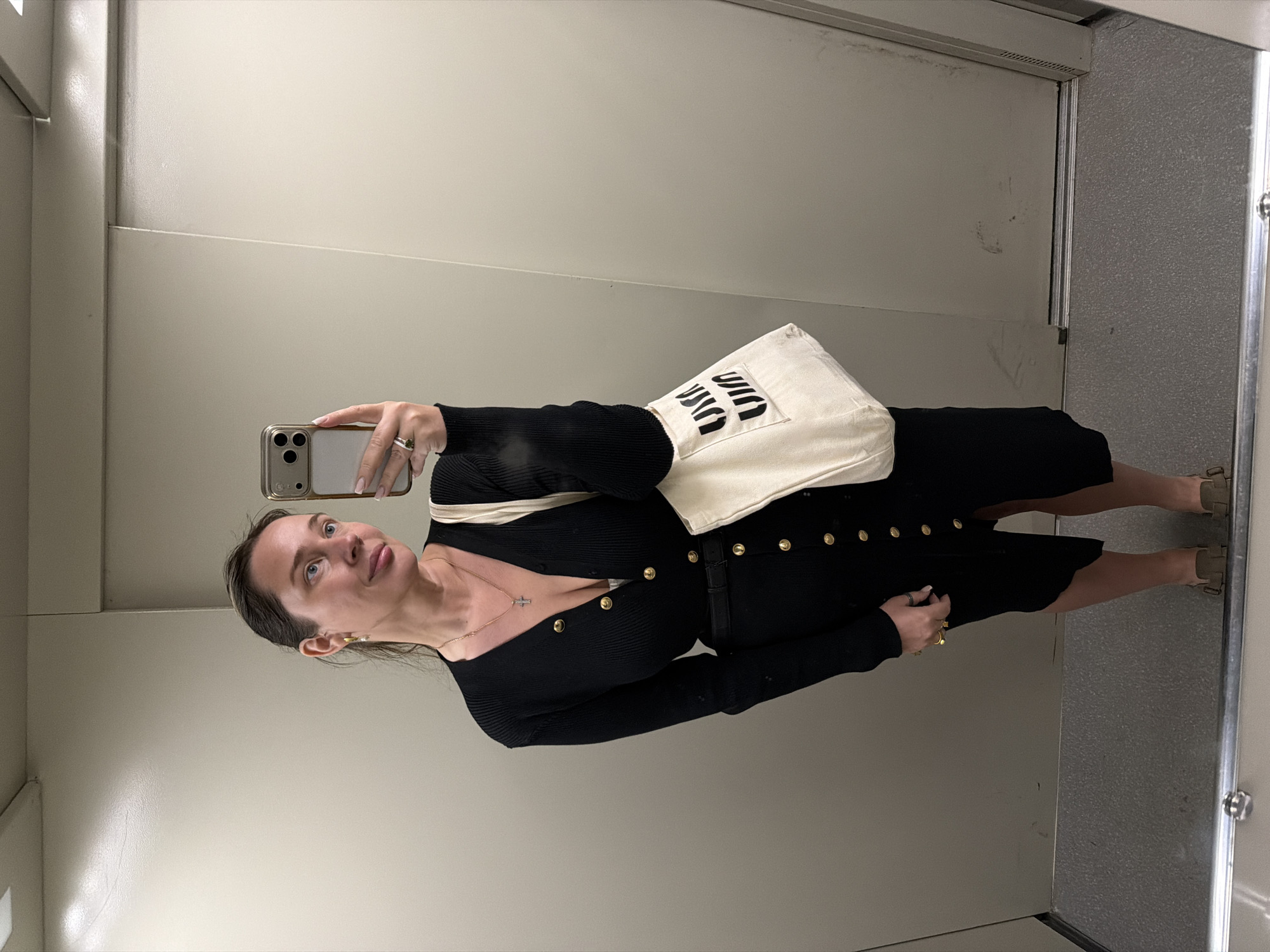 A woman stands in what appears to be a utilitarian elevator with beige walls and mirrored surfaces. She is holding a smartphone to take a picture of herself. She is wearing a black, button-down dress with gold buttons and a light-colored shoulder bag. The woman is also wearing a cross necklace. The location in the city of Chaikovsky, Russia cannot be verified.