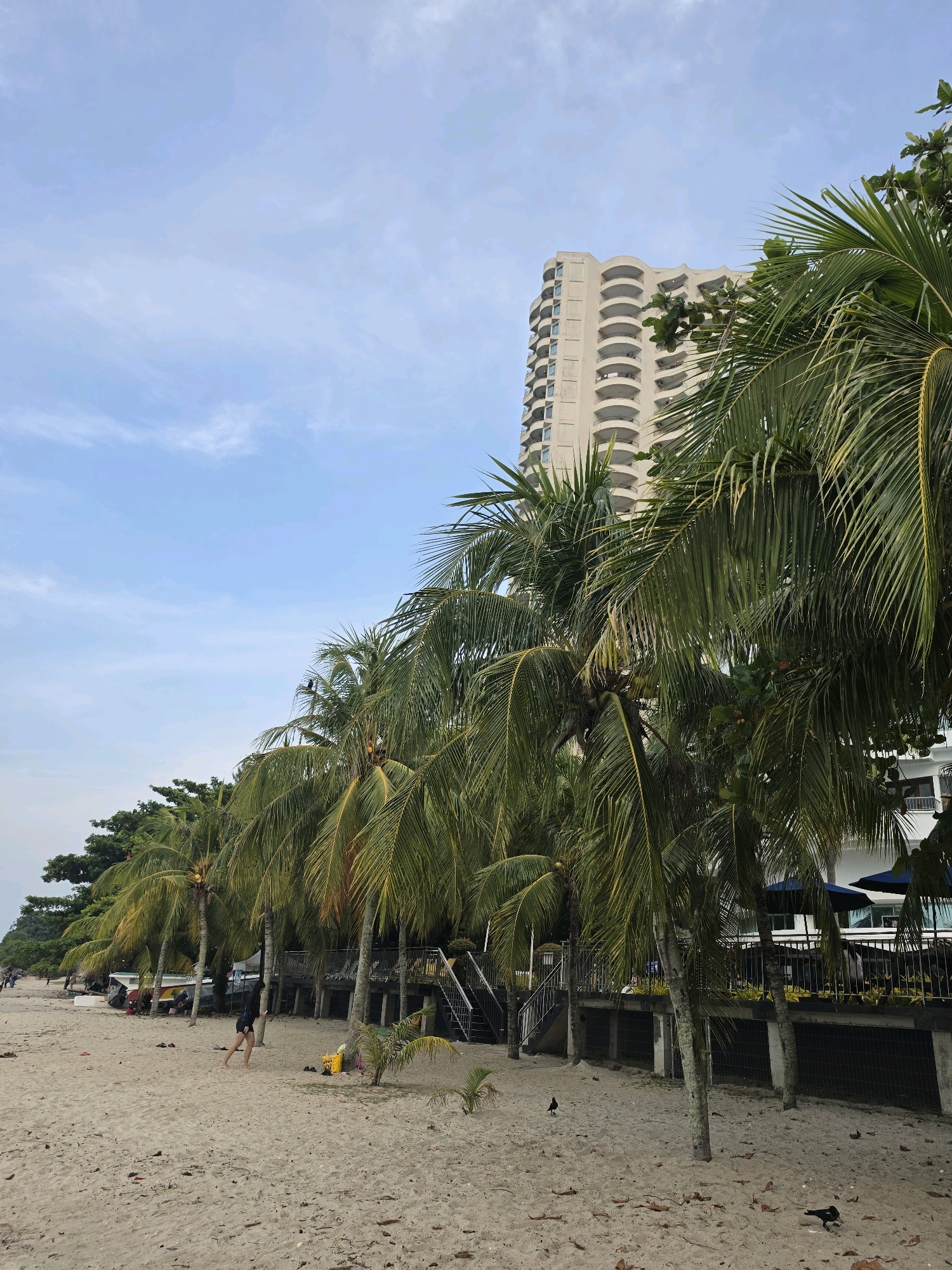 This is a photograph of a beach scene in George Town, Malaysia. In the foreground, there is a sandy beach with a few leaves scattered on it. A person is walking on the beach, and there is a yellow bucket and some shoes near a palm tree. In the midground, a line of palm trees runs along the edge of the beach, with a building and several umbrellas visible behind them. The building is a multi-story structure with balconies. In the background, the sky is partly cloudy. The overall impression is one of a tropical beach resort.