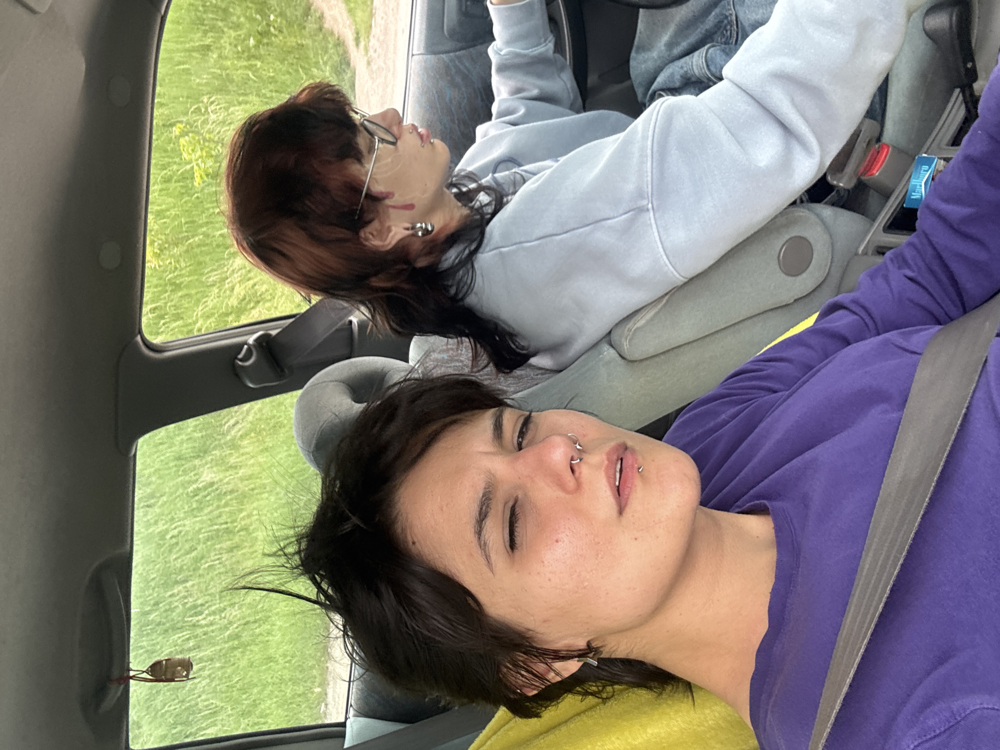 Two people are inside a vehicle. The driver has brown and black hair, glasses, and red marks on their face. They are wearing a light blue sweatshirt. The passenger has short, dark hair, piercings on their nose and lip, and is wearing a purple shirt. Both are wearing seatbelts. The background shows green grass and a road. It is impossible to infer the location in the city of Sofia.