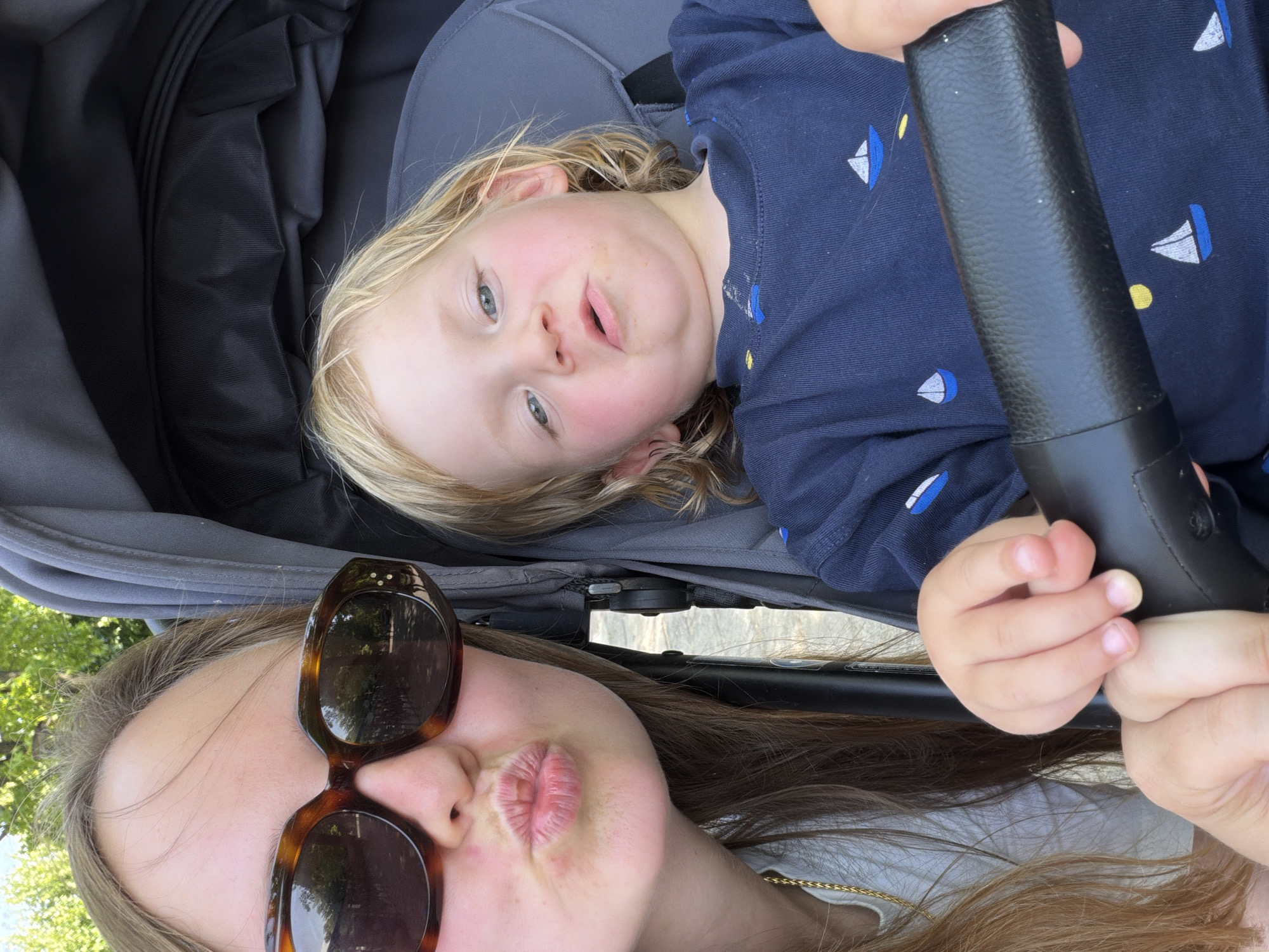 This is a selfie taken in the city of Como, Italy. A woman with long brown hair wearing large tortoiseshell sunglasses is leaning into the frame from the left, puckering her lips. She is wearing a white top with a gold chain. Beside her, in a stroller, is a young child with blonde hair, wearing a navy blue shirt with small sailboat patterns. The child is looking towards the camera with a slightly grumpy expression. The background shows greenery and a stroller.