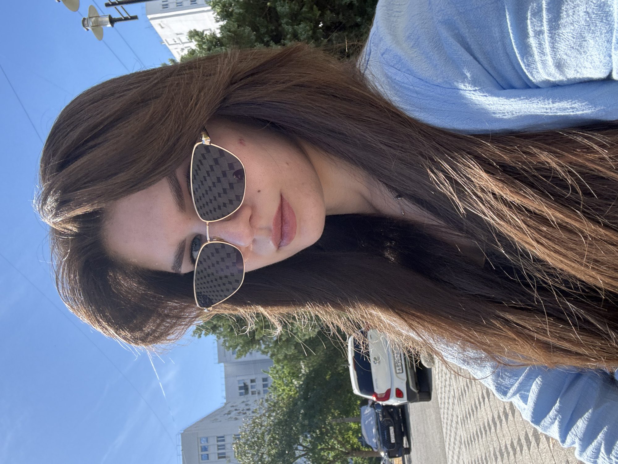 A young woman with long, brown hair is taking a selfie outdoors in what appears to be a city. She is wearing sunglasses with a gold frame and a light blue textured shirt. In the background, there are apartment buildings, trees, streetlights, and parked cars. The sky is clear and blue.