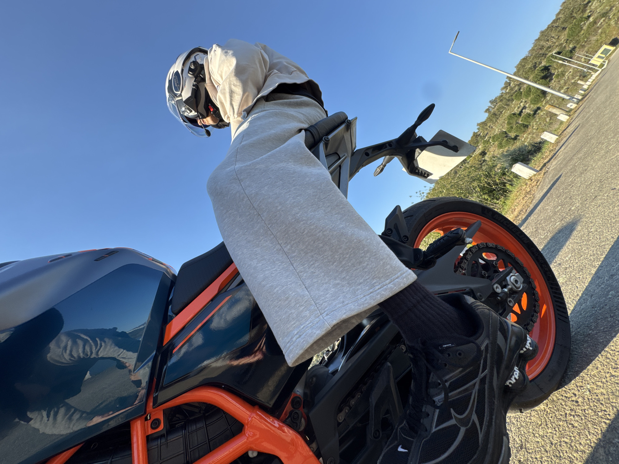 A low-angle shot shows a person sitting on a motorcycle. The person is wearing a helmet, a light-colored hoodie, gray sweatpants, black socks, and black sneakers. The motorcycle has a black and blue gas tank with orange accents. It has an orange wheel rim. The background includes a clear blue sky, a paved road, a grassy area, and a hillside with sparse vegetation. There is a lamppost and other roadside structures visible in the distance. It is difficult to pinpoint the exact location in Mirandela, Portugal, without more information.