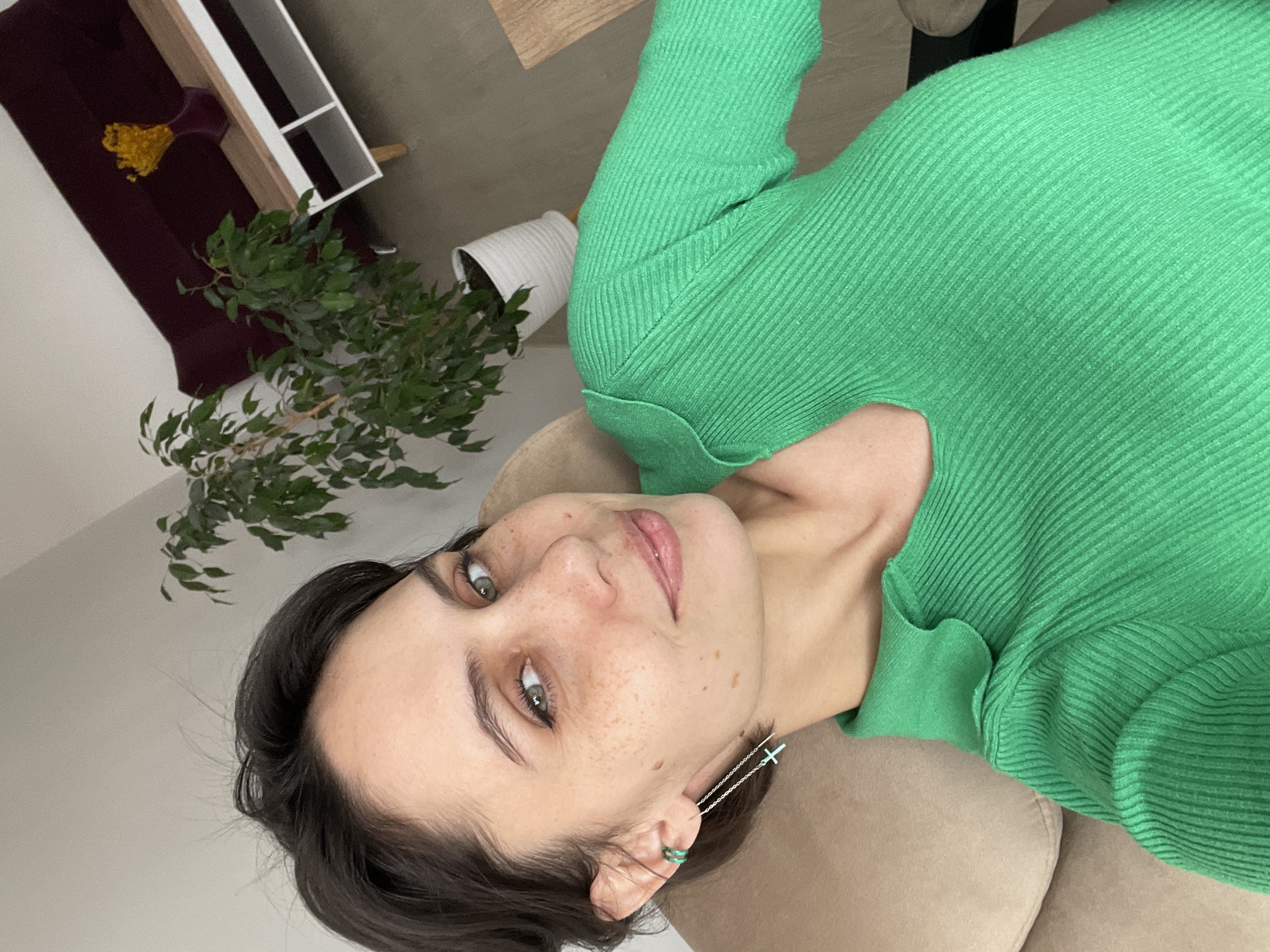 A young woman is depicted in a rotated, eye-level indoor shot, looking directly at the camera with a calm, soft expression. Her dark hair is swept back, revealing her face which features green eyes and numerous freckles across her nose and cheeks. She is wearing a form-fitting, bright green ribbed long-sleeved top. Her accessories include a silver chain earring with two small teal cross charms on her right ear, and a double teal ear cuff on her left ear.
She appears to be lying on a light beige upholstered sofa or armchair, which fills a significant portion of the frame. In the background, the setting reveals elements of a modern living space. To her left (as if the image were upright), a large green leafy plant in a white pot stands on the floor. Further back and to her right, there is a dark reddish-brown sofa with a small yellow floral arrangement in a dark vase, positioned next to a light wooden console table or shelf unit with white compartments. The walls are a plain light color. The scene is brightly and evenly lit, suggesting daytime. No text is visible in the image. The location is Izhevsk, Russia.