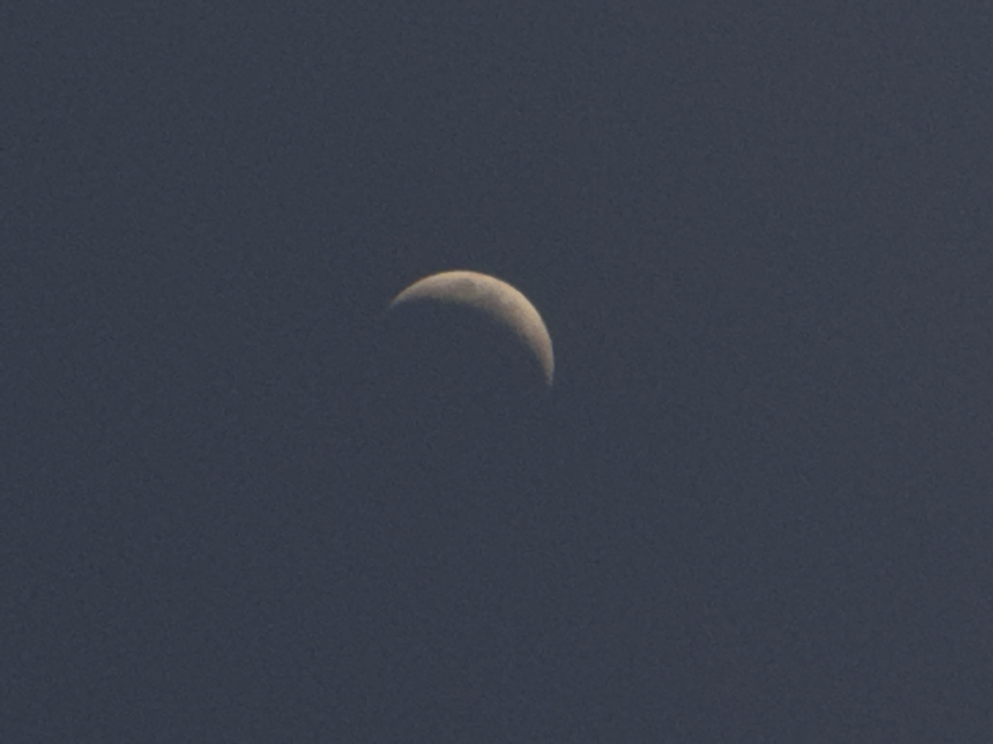 The image shows a crescent moon against a dark, slightly grainy sky. The moon is off-center, positioned slightly above the middle of the frame. The sky is uniformly dark with no other visible stars or celestial objects. There is no indication of the location or city of Mayak, Russia, visible in the image.