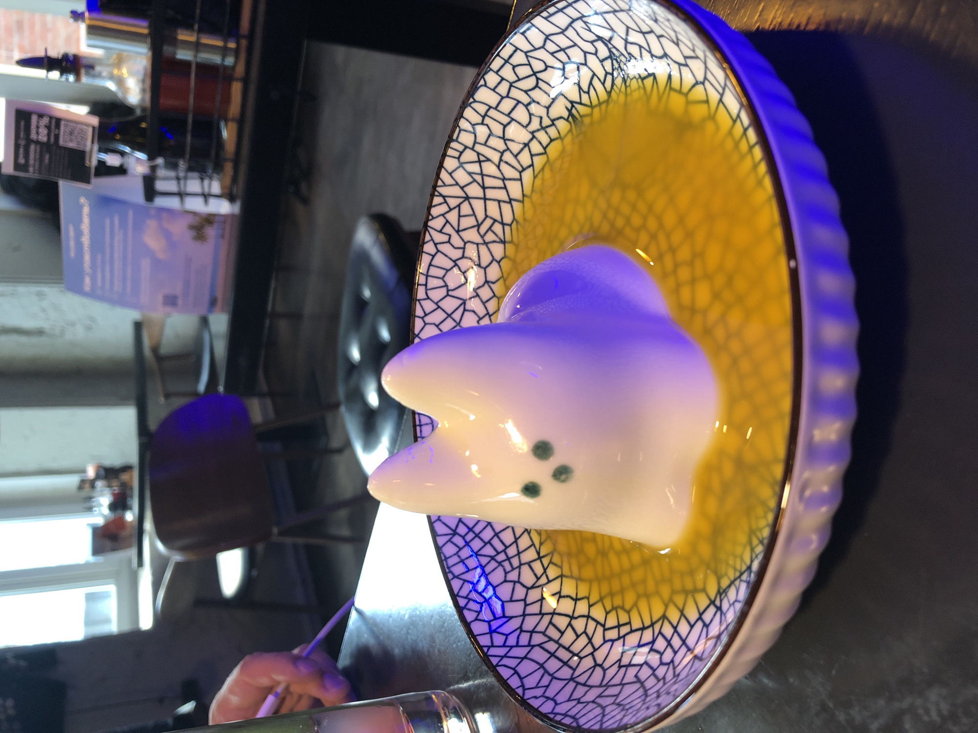 In Izhevsk, Russia, a white, cat-shaped dessert with three green dots for eyes is presented in a yellow liquid within a decorative bowl. The bowl features a black, intricate web-like pattern on a white background. The dessert is illuminated by purple and blue lights, casting reflections on the bowl's surface. The background is blurred, showing a table, chairs, and a window with natural light. A person's hand, holding a straw, is visible in the upper left corner, reaching towards the dessert.