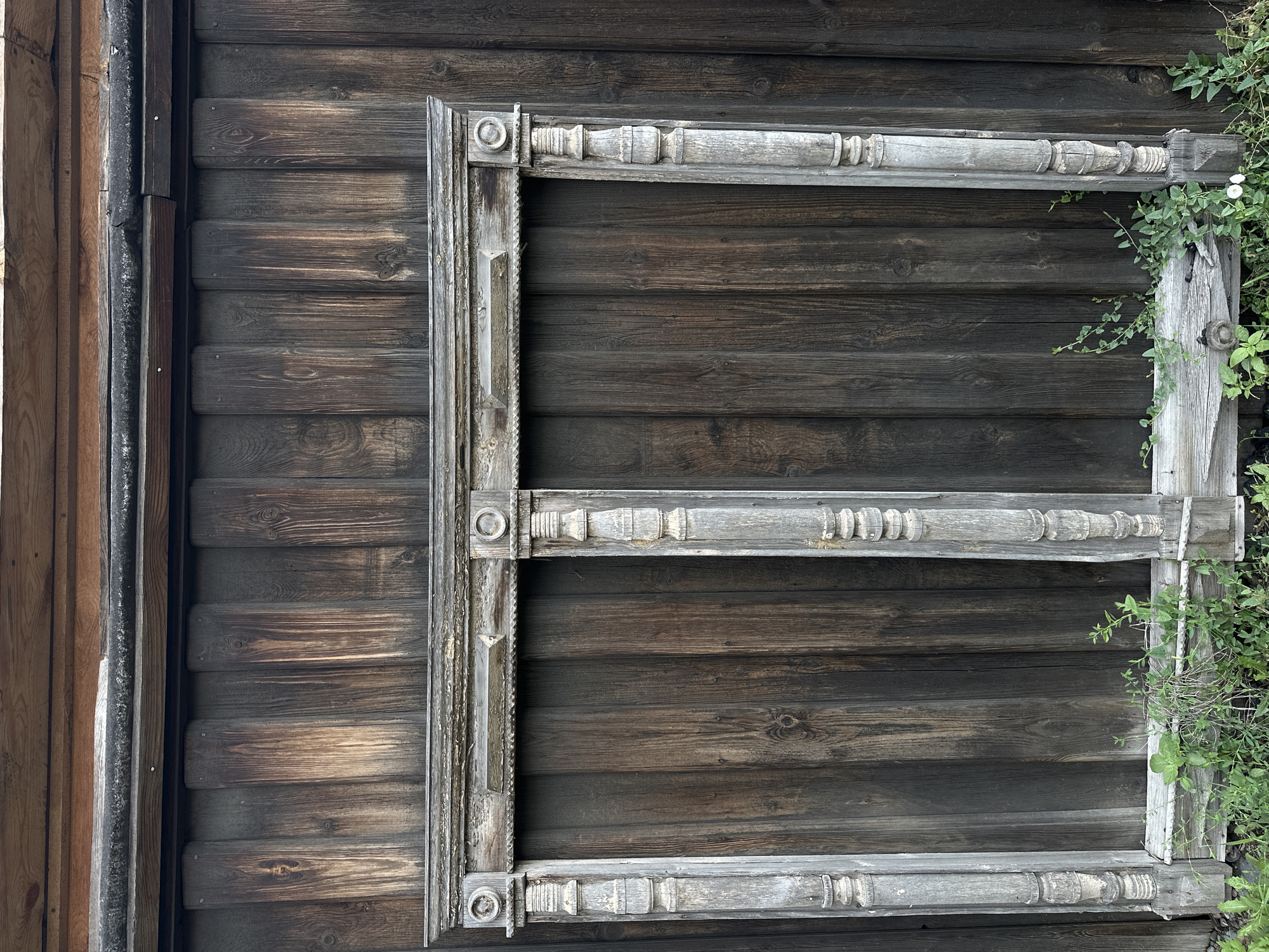 An aged, weathered wooden frame, likely for a double window, leans against a vertical wooden wall in the city of Nogornyi, Russia. The frame, painted a faded grey, shows signs of peeling paint and wear. It is ornately carved with decorative elements, including circular motifs at the top corners and stacked cylindrical shapes along the vertical posts. The wooden wall behind it is constructed of dark, rough-hewn planks, showing a distinct grain pattern and varied tones of brown and grey, suggesting it has also been exposed to the elements for a considerable time. Greenery, including grass and small vines, grows at the base of the wall and around the lower portion of the frame. The overall scene conveys a sense of age and abandonment.