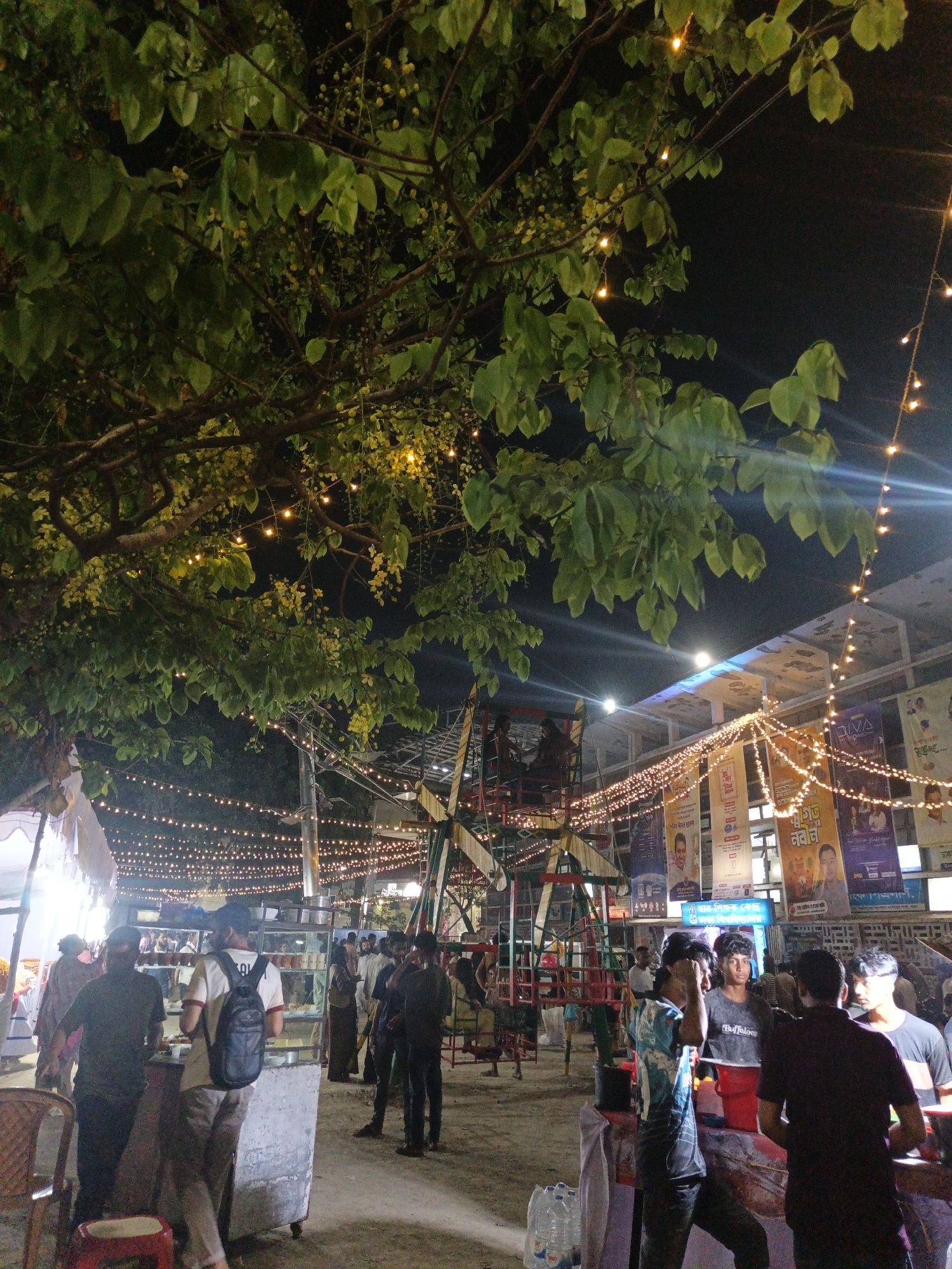 This nighttime photograph captures a lively outdoor gathering or market in Dhaka, Bangladesh, illuminated by numerous strings of warm fairy lights draped across the scene. A large tree with abundant green foliage and small yellow flowers (likely *Cassia fistula*) frames the upper left and center of the image, its branches intertwined with some of the lights.
Numerous people, mostly men, are scattered throughout the setting, socializing and interacting. In the foreground on the left, a man in a black t-shirt and another in a white t-shirt with a black backpack stand near a vendor cart, possibly selling refreshments or food from its glass-fronted display. Towards the right, a group of young men is gathered, with one wearing a t-shirt featuring the word "BUFFALOES."
In the mid-ground, a colorful, somewhat rustic carnival ride, resembling a small Ferris wheel or swing ride, adds to the festive ambiance. The ground appears to be unpaved dirt or uneven pavement. In the background, a building is adorned with multiple large, vertical banners. These banners display Bengali script and images of people, indicating they are likely advertisements, political promotions, or event announcements. One banner clearly features the English word "Diva." The overall scene depicts a bustling, social event under the artificial glow of the evening lights.