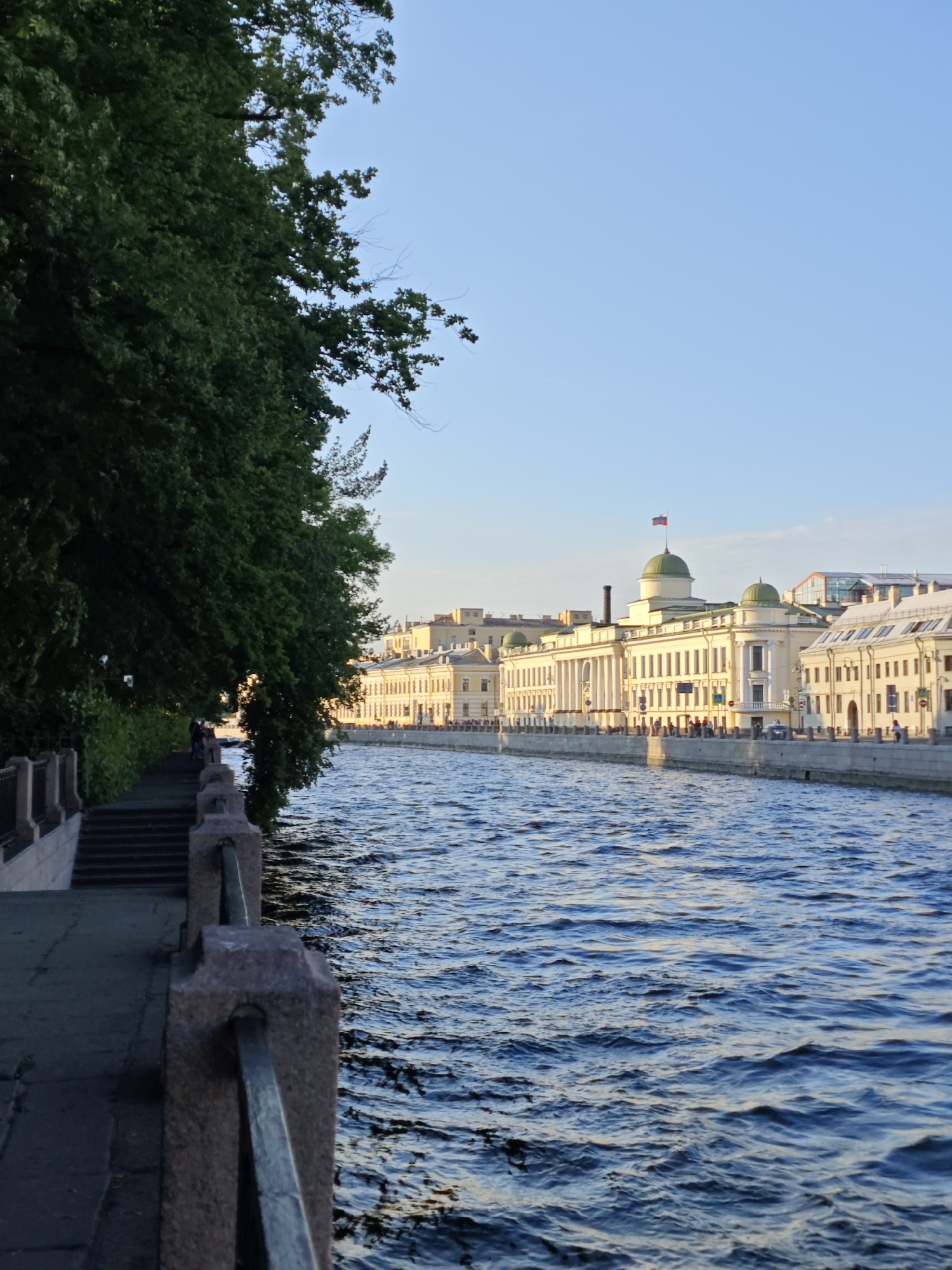 This is an eye-level, medium shot of the Fontanka River in Saint Petersburg, Russia, on a clear day. On the left, a tall, dense green tree dominates the foreground, its branches reaching towards the sky and partially obscuring the view of the opposite bank. A stone embankment with stairs leads down to the water's edge, bordered by a dark gray paved walkway and a stone railing. A few people are visible on the walkway. The river is a vibrant blue, with small waves reflecting the sunlight. Across the river, a row of grand, pale yellow buildings with white trim lines the embankment. The architecture features classical elements like columns and arched windows. One building is topped with a green dome and flies a Russian flag. The sky is a clear, light blue. The overall impression is one of a serene urban landscape on a sunny afternoon.