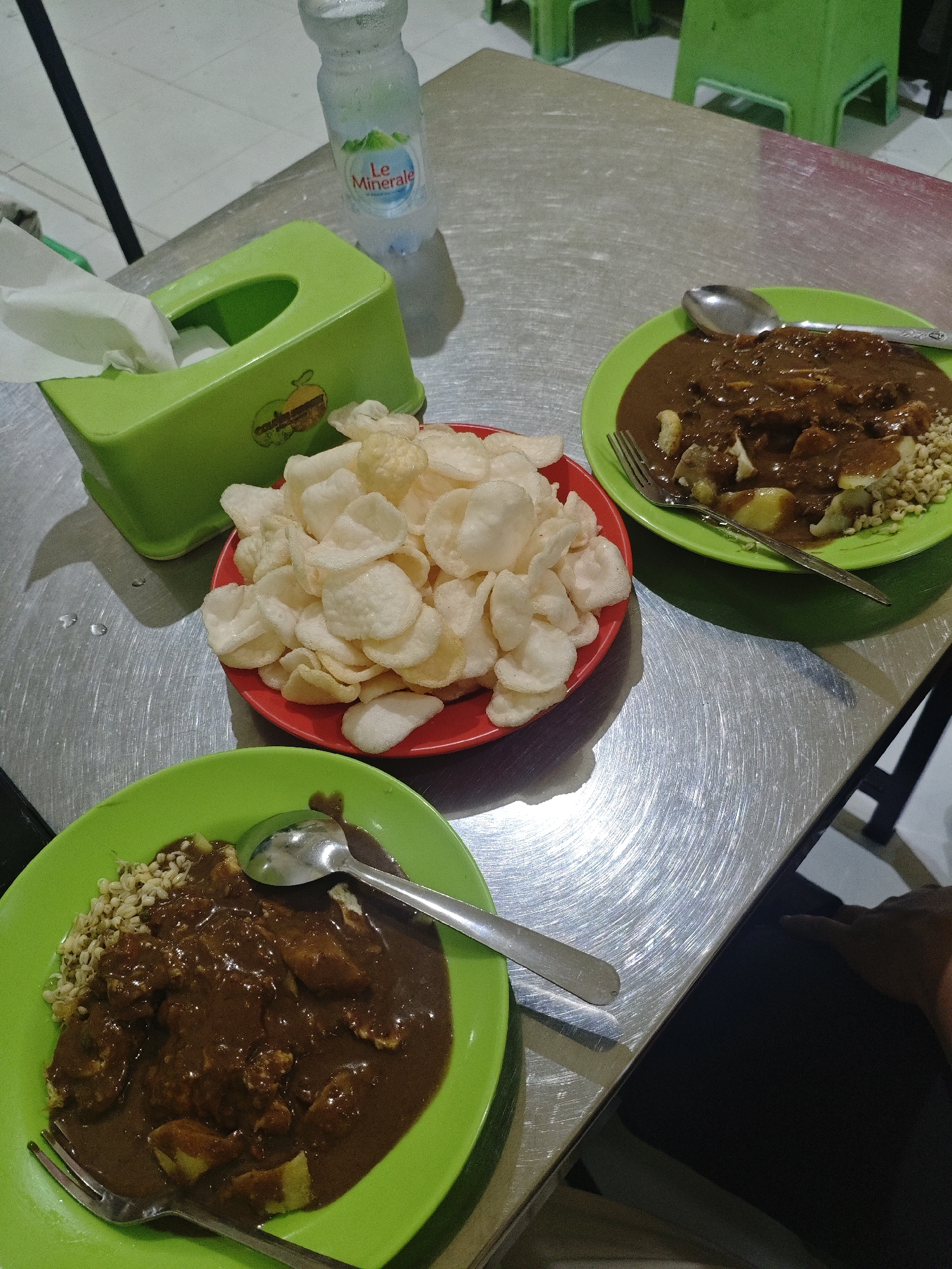 The image depicts a casual dining scene on a metallic table, likely in a street-side eatery or a local warung in Surabaya, Indonesia. Two bright green plates are filled with a dish featuring pieces of food in a dark, rich sauce, accompanied by a serving of bean sprouts. One plate also has a pile of white crackers. A red bowl holds a generous portion of these crackers. A green tissue box, partially filled with white tissues, sits to the left of the food. A bottle of "Le Minerale" drinking water is positioned behind the tissue box. A spoon rests on one of the green plates, and a fork is placed on another. The overall ambiance suggests a simple and unpretentious meal.
