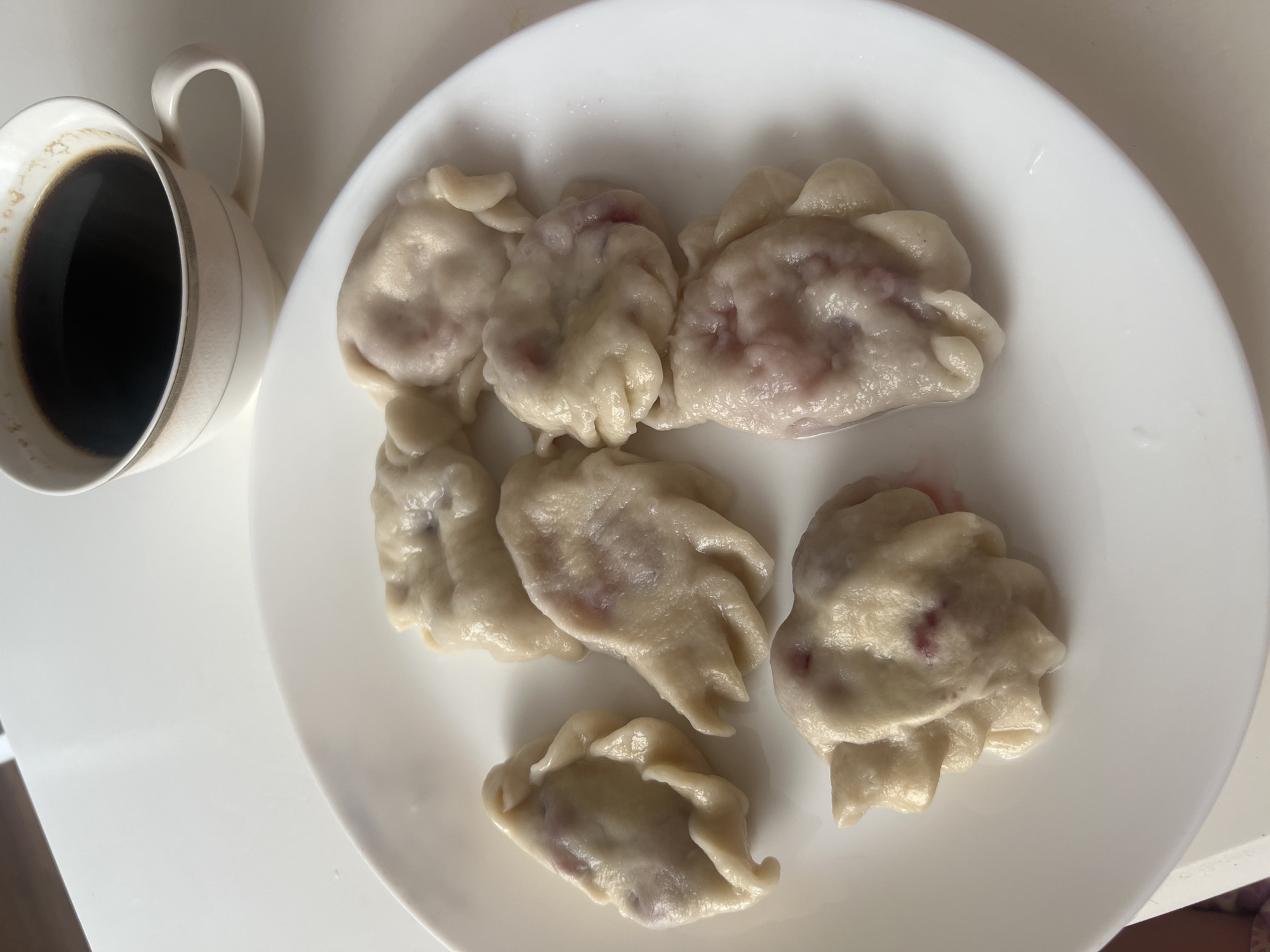 A white plate with six pierogi filled with what appears to be berries sits on a white surface. To the upper right of the plate is a white mug filled with black coffee. The scene appears to be indoors, likely a domestic setting. There is no visible indication of the city of Lipetsk, Russia.