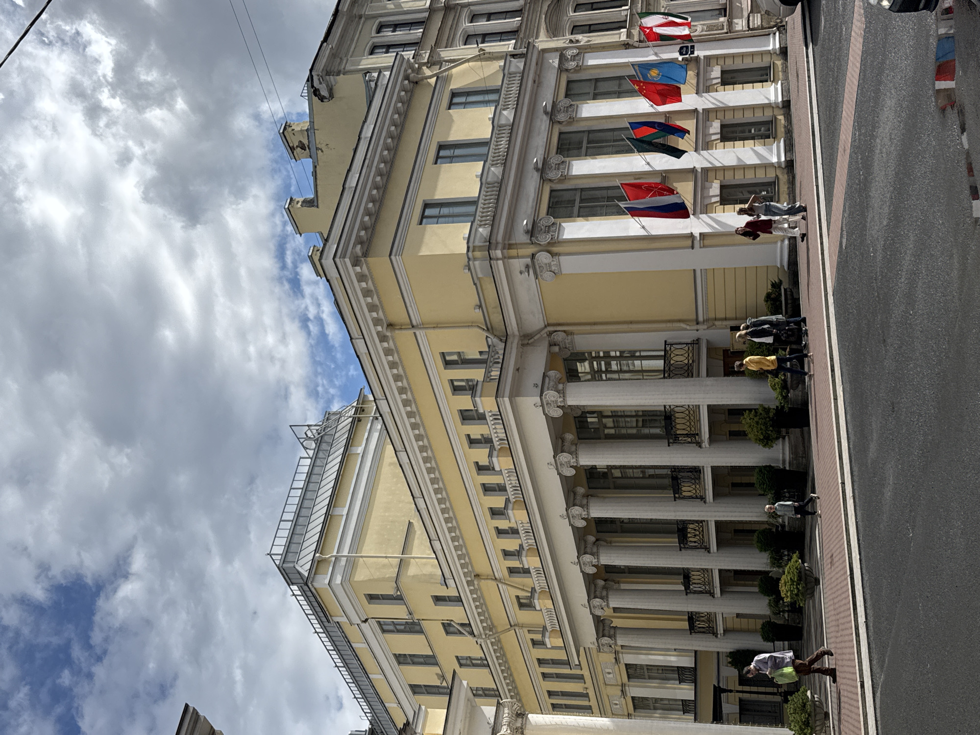 The image shows the yellow facade of a neoclassical building in Saint Petersburg, Russia, under a cloudy sky. The building features a colonnade with Corinthian columns on the ground floor and a balustrade on the upper floor. Several flags are displayed in front of the building, including the flags of Russia, Kazakhstan, and China. People are walking on the sidewalk in front of the building. One person is walking with a green bag, another is walking with a backpack, and a group of three young women are walking together. A child is also walking on the sidewalk. The street in front of the building is paved with asphalt and has a red and white striped crosswalk. The reflection of the flags is visible in a puddle on the street. The architectural style and the presence of flags suggest this building is a significant institution, possibly a consulate or embassy, located in the historic center of Saint Petersburg.