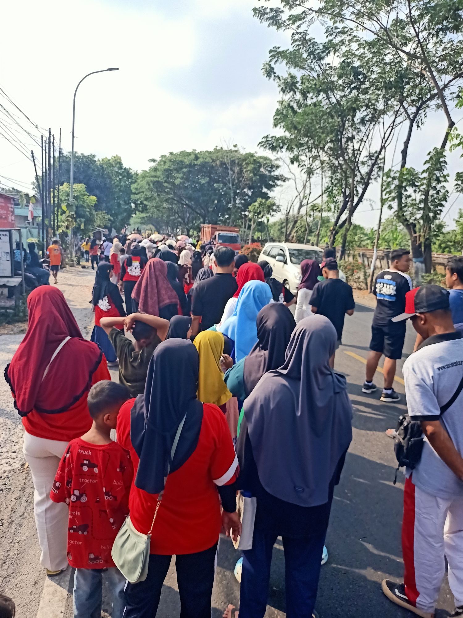 A group of people are walking on the side of a road in Kendung, Indonesia. The scene is outdoors with a bright sky and trees lining the road. The people are dressed in casual clothing, with many wearing headscarves. Some are walking in the same direction, suggesting a procession or a gathering. There are vehicles, including a white car and a truck, visible on the road. The road surface appears to be asphalt, and there is vegetation and a lamppost visible along the roadside. The overall impression is of a community event or activity taking place in a semi-urban or suburban environment.