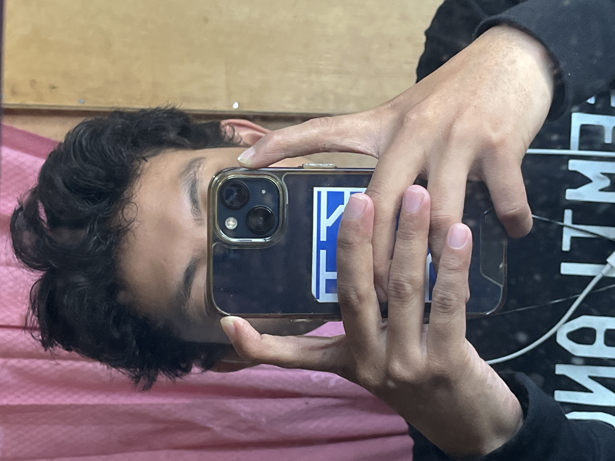 A person is taking a mirror selfie. They are holding a smartphone up to their face, obscuring most of their features. The phone has a clear case with a blue and white sticker on the back. The person has dark, curly hair and is wearing a black hoodie. The background consists of a pink curtain and a light brown wall. The image was taken in the city of Sejahtera, Indonesia.