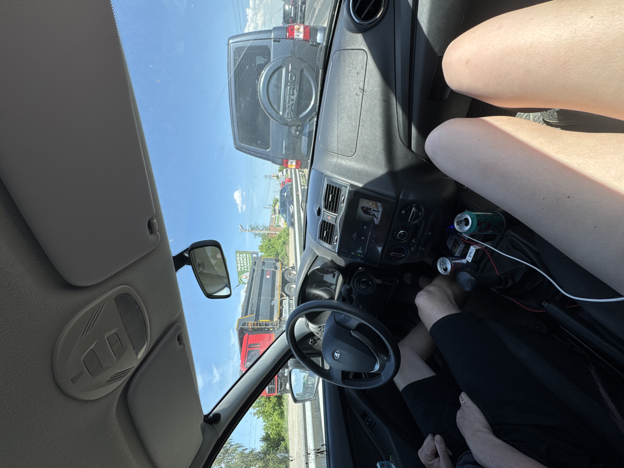 The image shows the interior of a car, specifically the driver's side. A black "Patriot" SUV is directly in front of the car, visible through the windshield. The driver's legs are visible in shorts and flip-flops, and a passenger is visible in the center console. The car is stopped in traffic, as evidenced by the other vehicles in the distance and the fact that the driver's foot is not on the pedals. A sign in the distance appears to be written in Russian.