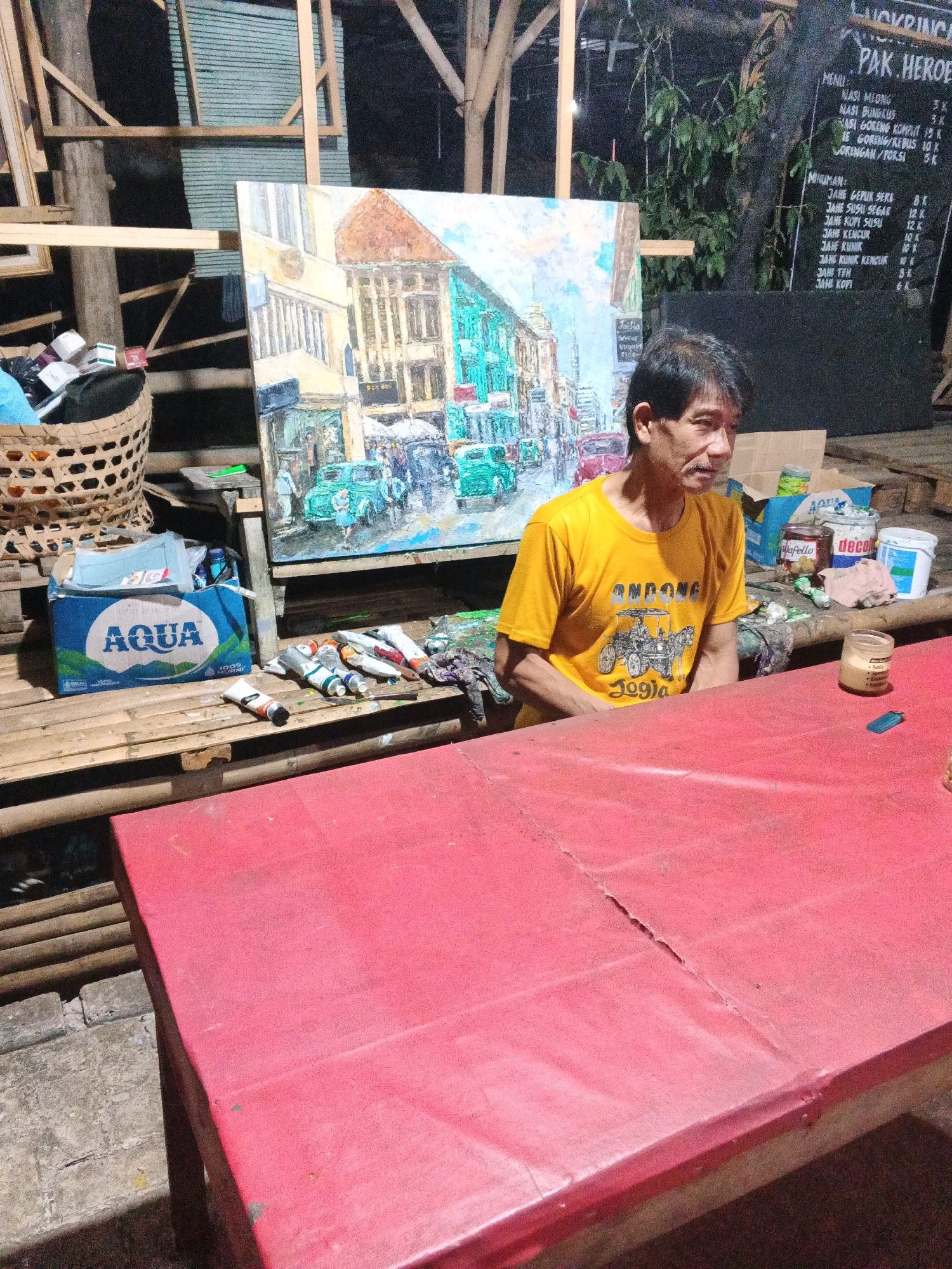 The image depicts a man sitting at a red table in what appears to be an outdoor setting, possibly a workshop or an artist's space, at night. The man, who is the central subject, is wearing a yellow t-shirt with an illustration and text that includes "JOGJA". He is looking to his right, away from the camera.
Behind the man, an unfinished painting of a street scene with buildings and vintage cars is displayed on an easel. To the left of the painting, a woven basket is filled with various items, including what look like cigarette packs. A blue "AQUA" water bottle is also visible. Several tubes of paint are scattered on a wooden surface in front of the painting. In the background, a blackboard menu with Indonesian writing lists food and drink items, suggesting the location might be a food stall or restaurant. The overall environment is dimly lit, with wooden structures and some foliage visible.
While the image shows elements that could be associated with a public space, there is no specific visual information within the image itself that confirms its exact location in Ungaran, Indonesia. The menu and clothing with Indonesian text indicate a location within Indonesia, and "JOGJA" on the t-shirt refers to Yogyakarta, but the precise setting is not explicitly identified as Ungaran.
