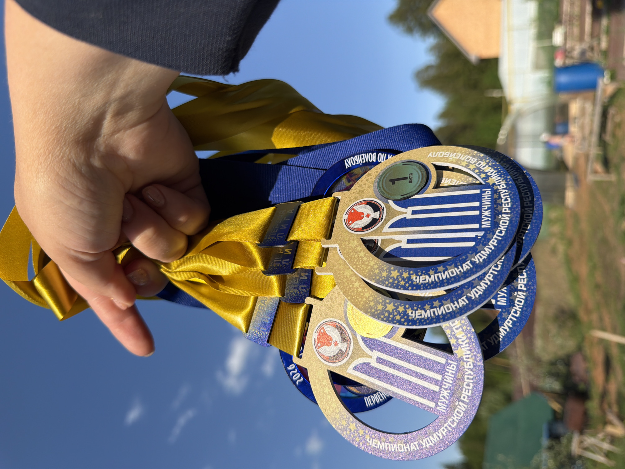 A person's hand holds several gold medals with ribbons in an outdoor setting. The medals are circular with a blue and gold design and Russian text, indicating they are from a "Championship of the Udmurt Republic" competition. One medal has a sticker indicating "1st place." The ribbons are gold and blue. The background shows a blue sky, trees, and possibly a greenhouse or shed. Based on the presence of "Chaikovsky" on the medals, the location might be the city of Chaikovsky, Russia.