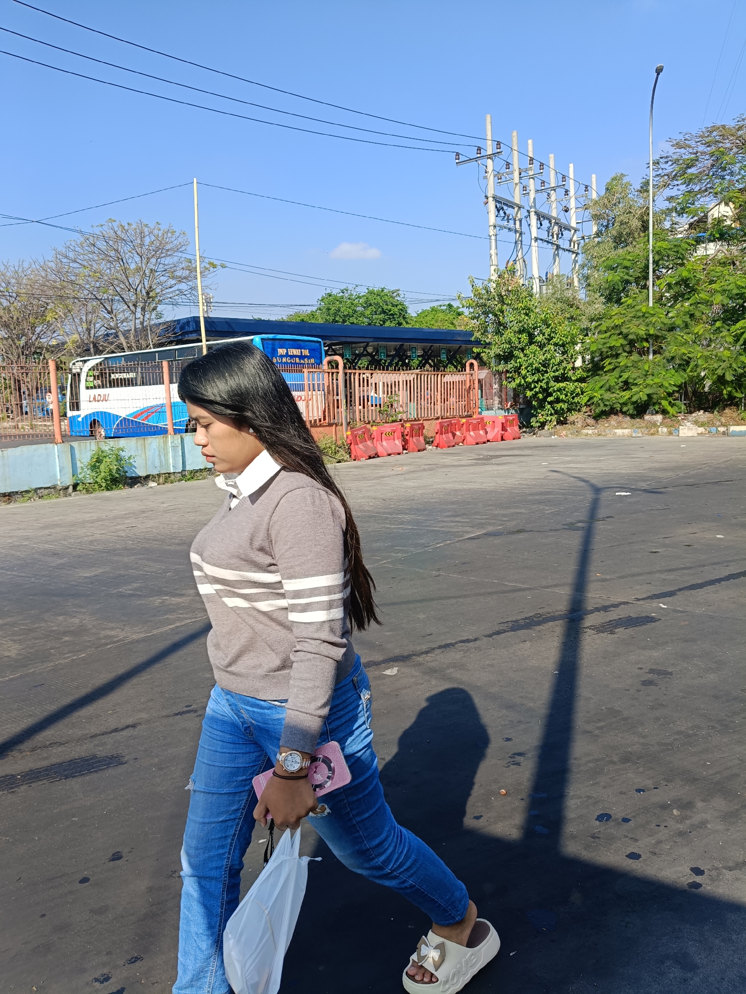 In Kedungrejo, Indonesia, a young woman with long, dark hair walks across an asphalt surface, likely a road or parking lot, under a clear blue sky. She is wearing a gray sweater with white stripes, blue jeans, and white platform sandals with a bow detail. She is holding a pink phone and a white bag in her right hand. To her left, a blue and white bus with the text "JMP LEWAT TOL" and "GUNGURASIH" is parked behind an orange metal fence. In the background, utility poles with electrical wires are visible, along with trees and a street light. The lighting suggests it is daytime, with shadows cast on the ground.