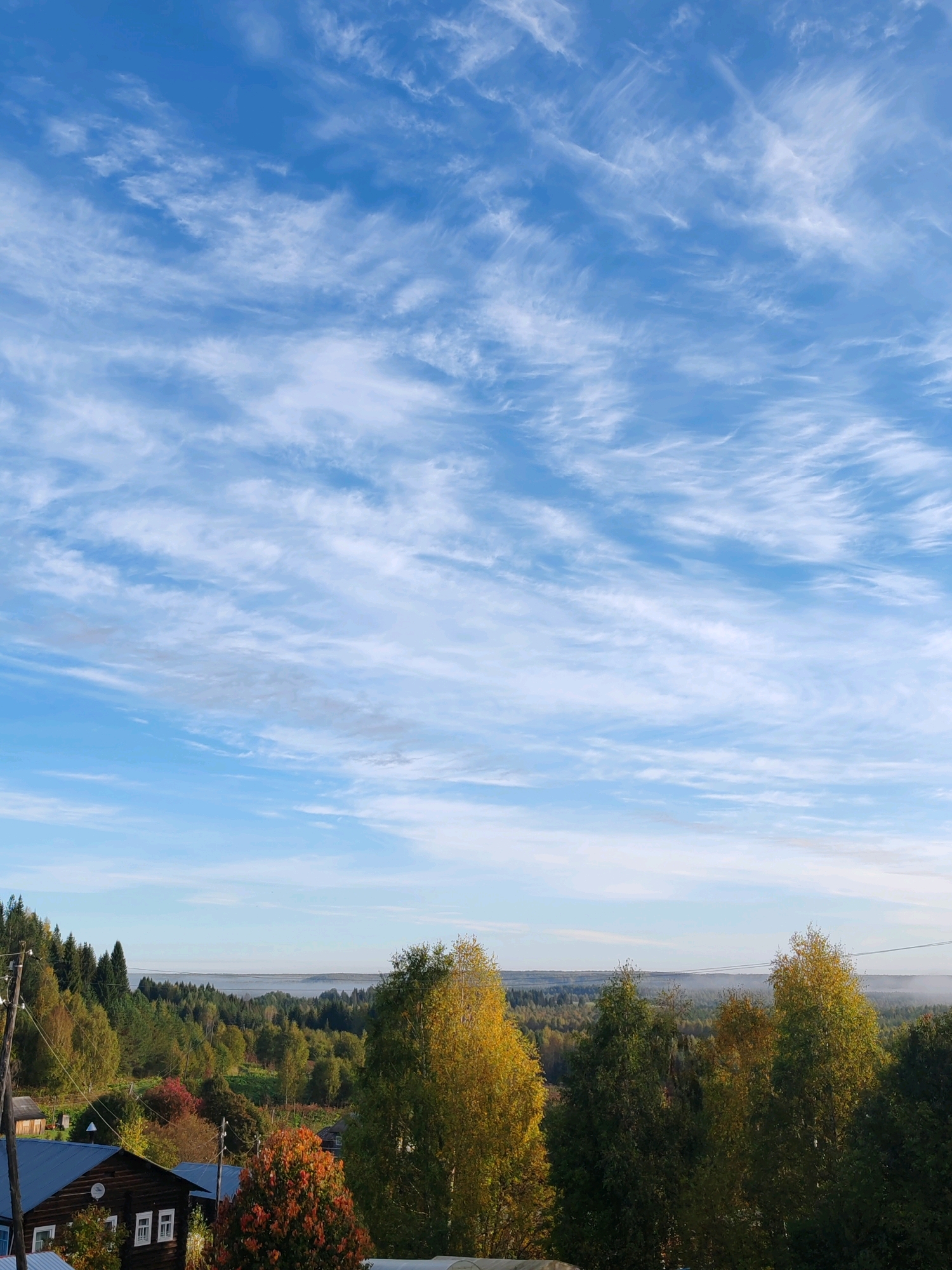 The image captures a panoramic view of a rural landscape in Chukhlem, Russia, likely during the autumn season. The upper portion of the image is dominated by a vast expanse of blue sky, streaked with wispy cirrus clouds that stretch horizontally across the frame. The lower half of the image reveals a rolling terrain covered in a dense forest. The foliage displays a vibrant mix of green, yellow, and red hues, indicative of autumn's changing colors. In the foreground, several wooden structures, typical of Russian rural architecture, are visible. One prominent building, a dark wooden house with white-framed windows, sits to the left. Another structure with a blue roof is partially visible. Utility poles with electrical wires are also present, suggesting the presence of a settlement. A thin layer of mist hovers over the distant tree line, adding a sense of depth and atmosphere to the scene. The overall impression is one of a serene and picturesque countryside.