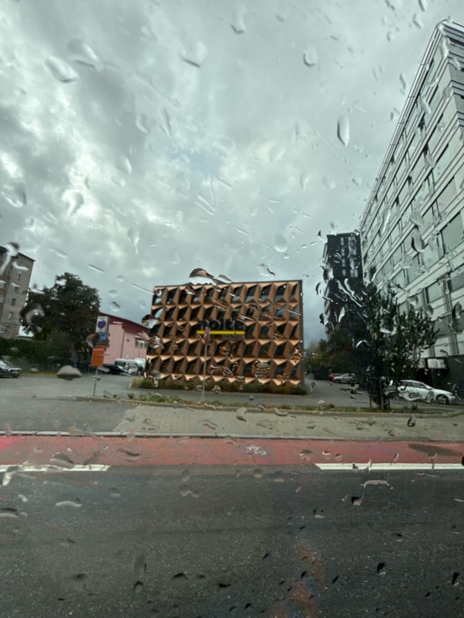 The image depicts a cloudy day in Tallinn, Estonia, with raindrops on the glass through which the scene is viewed. The foreground shows a wet asphalt road with red and white markings, likely a pedestrian crossing or bike lane. In the midground, there's a unique, modern building with a facade of triangular, copper-colored panels that catch the light. Adjacent to this building, a darker, taller structure is visible. To the left, a multi-story residential building and trees are present. A few cars are parked or driving on the street. The overall atmosphere is overcast and damp.