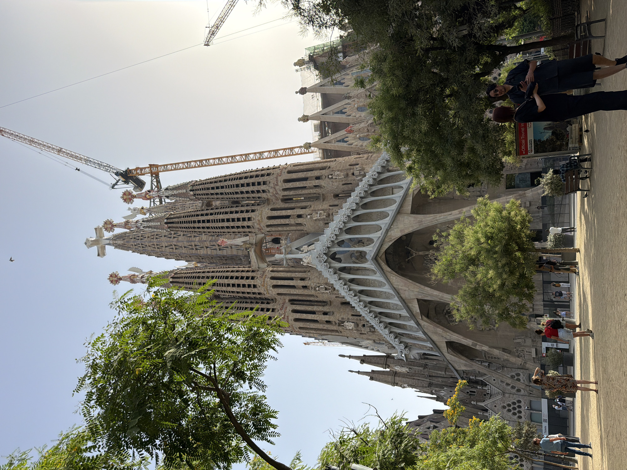 The image shows the Sagrada Familia basilica in Barcelona, Spain. The basilica is a UNESCO World Heritage Site and is still under construction. In the foreground, a group of people are standing in a park, some are looking at the basilica, while others are engaged in conversation. The basilica itself is a magnificent structure with intricate facades and soaring towers. Cranes are visible in the background, indicating ongoing construction work. The environment is sunny and clear, with trees providing shade and a park setting.