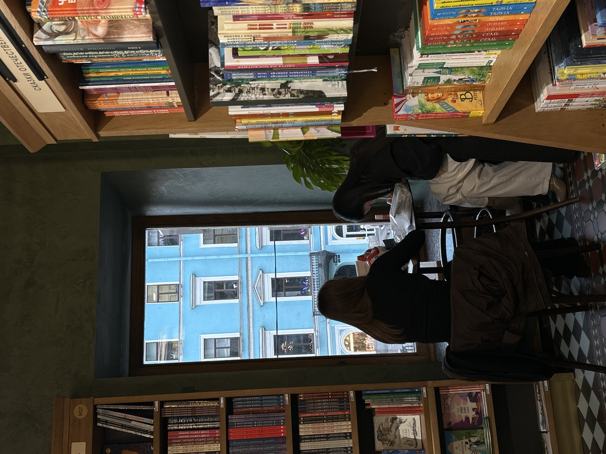 The image is taken from inside a building, likely a bookstore or cafe, in Saint Petersburg, Russia. The scene is dominated by a window providing a view of a blue building exterior. Two individuals are seated near the window, engaged in activities that suggest reading or working. One is wearing white pants and a dark jacket. The second person is wearing all black.
The environment is characterized by shelves filled with books. The books appear to be in Russian, with titles visible upon close inspection. The bookstore setting is highlighted by the presence of signs containing Russian text. The location is likely near the center of the city due to the presence of the bright blue building, which is near several famous sites.