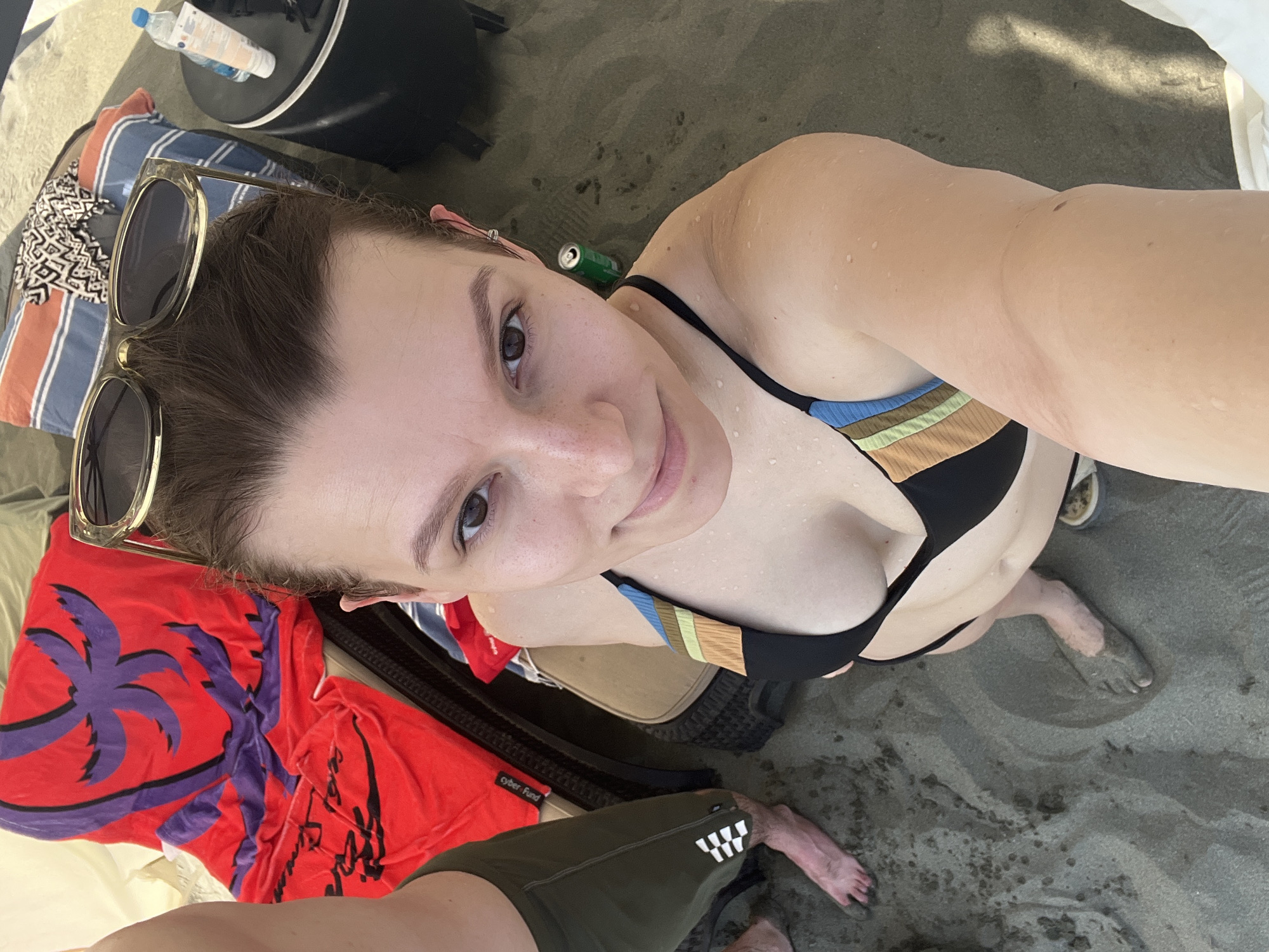 A woman lies on a sandy beach in Agios Tychona Community, Cyprus, taking a selfie with her phone. She is wearing a black bikini with a colorful striped pattern on the top. Sunglasses with light-colored frames rest on her head, and she has a pink clip in her hair. Beside her, a green soda can lies on the sand, and a red towel with a palm tree design is visible. She appears relaxed and possibly enjoying a sunny day, as suggested by the bright lighting and her attire. The background shows the sandy beach and hints of beach furniture. Partially visible in the foreground are a pair of dark green swim trunks and another person's feet, one with sand on it.