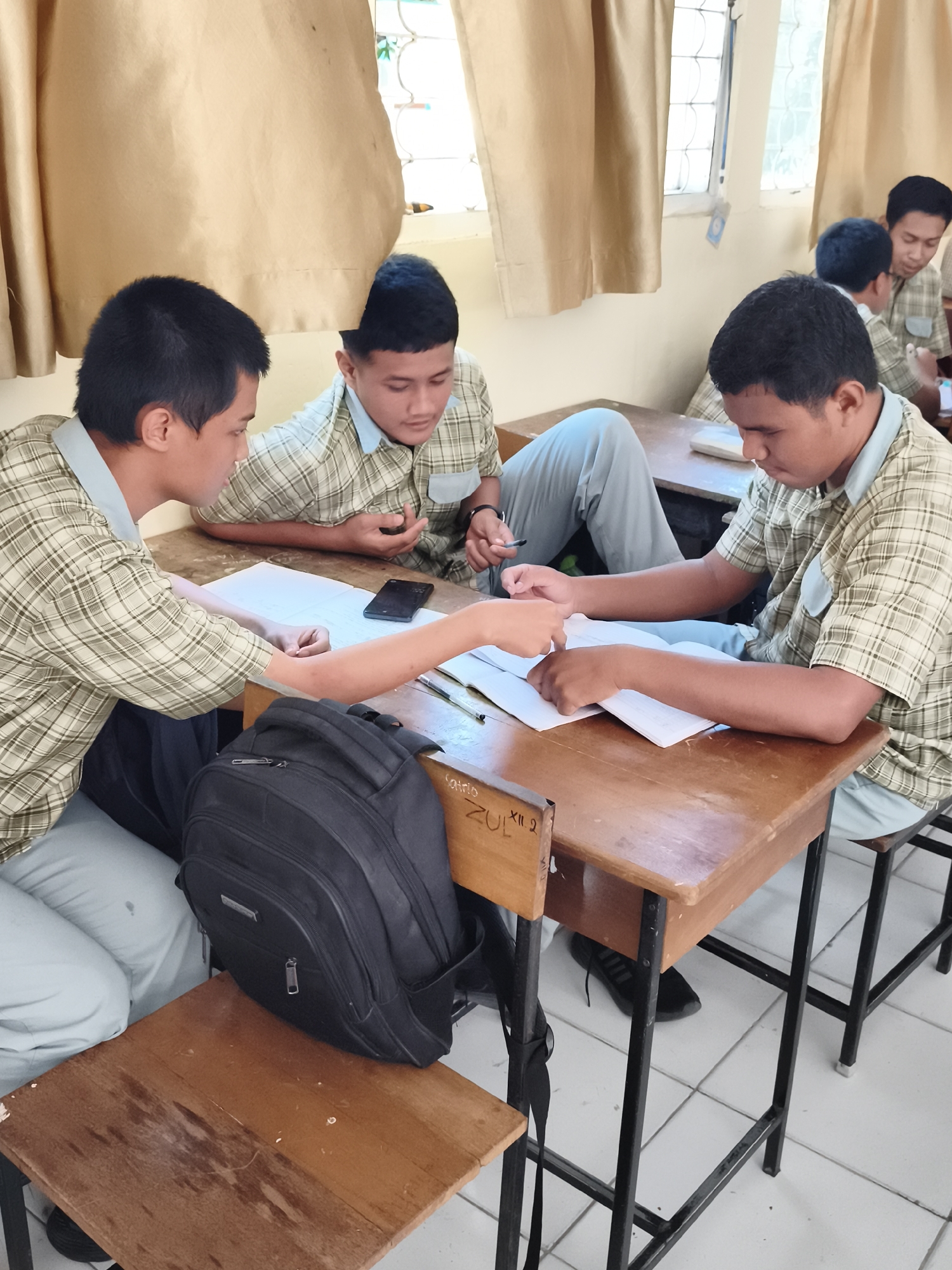 In Makassar, Indonesia, three male students wearing light green plaid shirts and light blue pants are seated at wooden desks in a classroom. The students are engaged in group study. One student in the foreground, facing left, has his hands on a book and appears to be pointing at it. Another student, in the center, is also looking at the book and holding a pen. The third student, in the background on the right, is looking down at a book and a mobile phone on the desk. A black backpack is placed on the floor beside the student on the left. The classroom has large windows covered by beige curtains, and the floor is tiled with white squares. The desks are worn, with some inscriptions visible on the left desk.