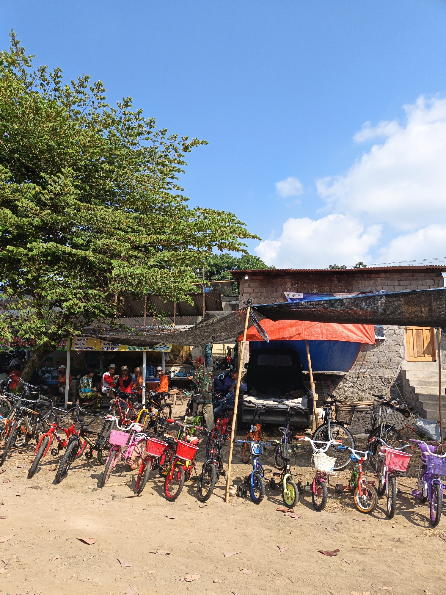 The image depicts an outdoor scene in Jatinom, Indonesia, under a clear blue sky with scattered white clouds. A large tree with dense green foliage dominates the left side of the frame, providing shade over a row of parked bicycles. In the background, a simple open-air structure appears to be a food stall or marketplace, with people sitting and interacting. The structure has a corrugated metal roof and brick walls. A variety of bicycles are parked in the foreground, ranging from adult-sized to children's models, many adorned with colorful baskets. The ground is covered with dry earth and fallen leaves, suggesting a rural or semi-urban setting. The overall impression is one of a casual, community gathering spot, likely a local market or roadside attraction.