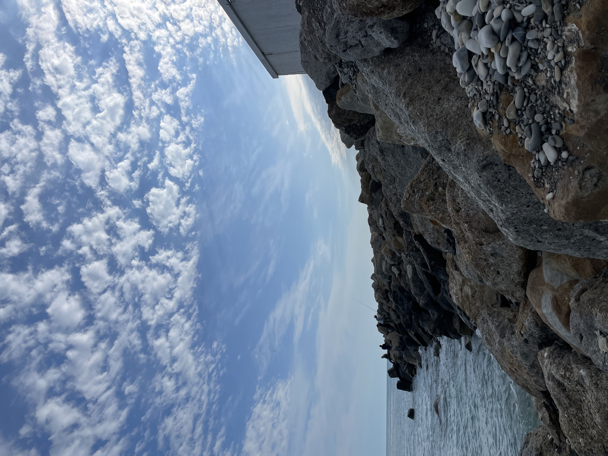 The image shows a view from the city of Olginka, Russia, looking out over the Black Sea. The sky is blue with scattered white clouds. A concrete structure and large rocks are visible on the right side of the image. People are standing on the rocks along the shoreline. The sea is calm and stretches to the horizon.