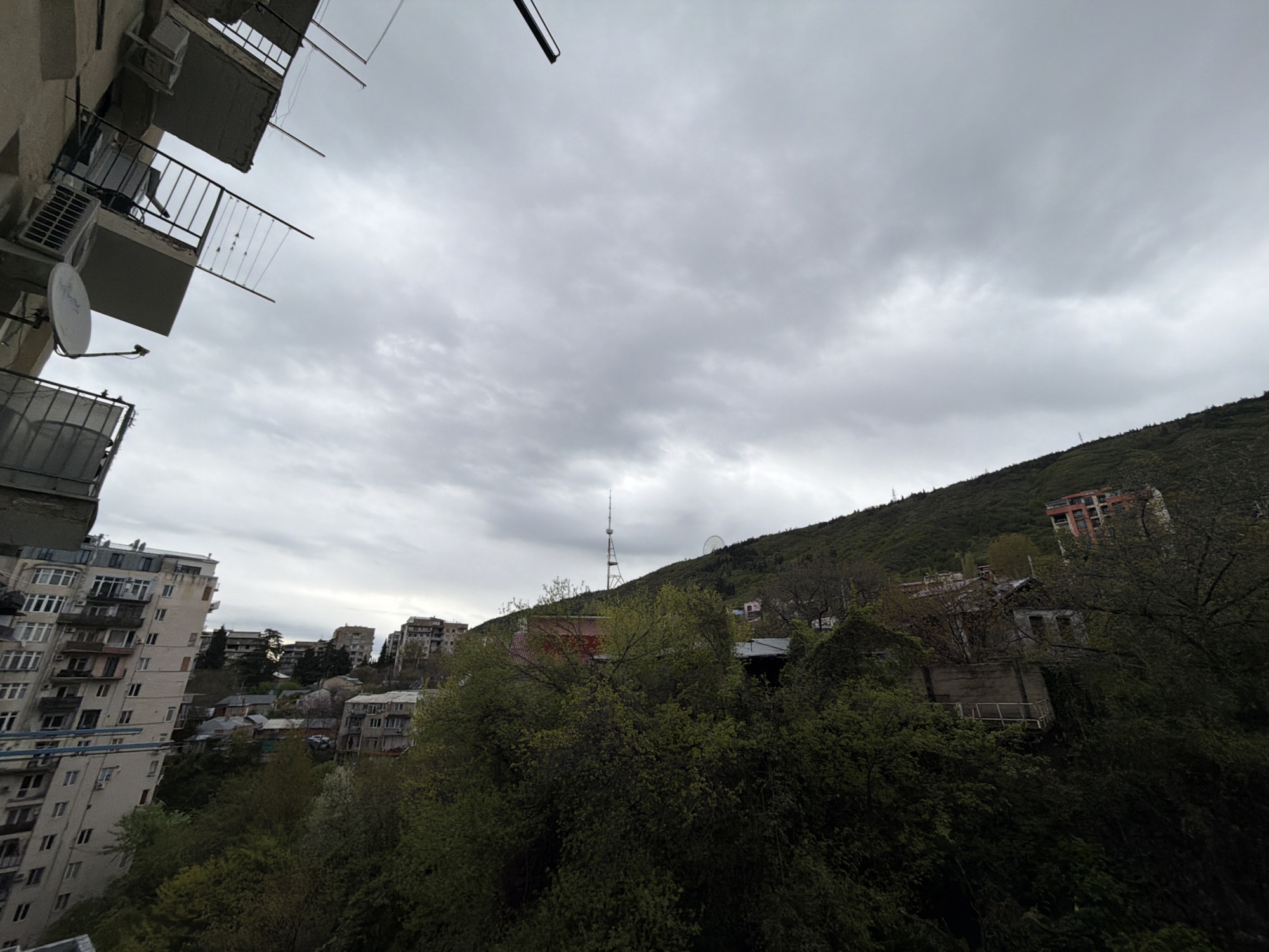 This image captures a panoramic view of a hillside city, specifically Tbilisi, Georgia, under an overcast sky. The foreground features the exterior of a building, likely where the photograph was taken from, showing parts of balconies with metal railings, several air conditioning units, and a white satellite dish.
Looking out from the building, the scene unfolds with a mix of urban structures and dense natural greenery. To the left, several multi-story apartment buildings with numerous windows and balconies are visible, characteristic of post-Soviet era architecture. The buildings appear somewhat densely packed, integrated into the hilly landscape.
The middle ground and background are dominated by a lush, green mountain or hill covered in trees and vegetation, suggesting either spring or early summer. Nestled within this greenery are a few scattered residential structures, some appearing older or partially obscured by foliage. Prominently rising from the hilltop is the slender, iconic Tbilisi TV Tower, a significant landmark. To the right of the TV Tower, a Ferris wheel can be partially seen, indicating the location of Mtatsminda Park.
The sky is uniformly grey and cloudy, suggesting an overcast day with muted light. No direct human activity is visible, but the presence of residential buildings implies a bustling city life. There is no visible text in the image. The overall atmosphere is calm and slightly subdued due to the cloudy weather, highlighting the unique topography of Tbilisi with its blend of urban development and natural hills.