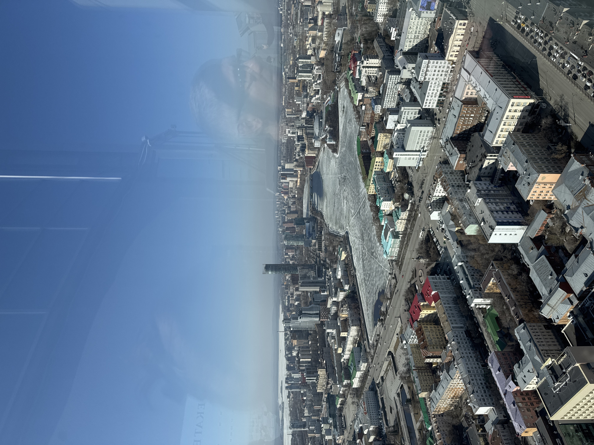 The image depicts an aerial view of Yekaterinburg, Russia, seen through a window, possibly from a high-rise building or observation deck. The city is predominantly composed of medium-rise buildings arranged along a street grid. A prominent feature is a frozen river, likely the Iset River, winding through the city center. The day is sunny, evidenced by the clear blue sky and bright reflections in the window. A tall building can be seen in the distance. The window shows a reflection of a person. A text is also reflected in the window.