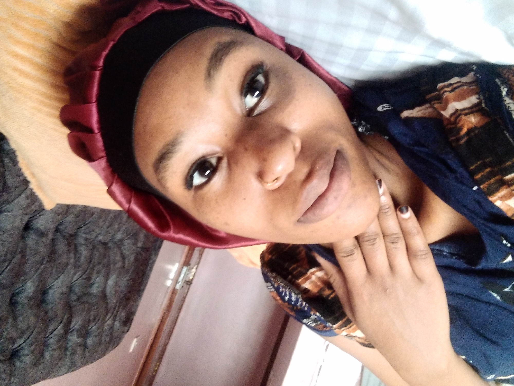 The image is a close-up, low-angle, selfie-style photograph of a young Black woman lying down indoors. The woman is wearing a navy blue top with a brown and black patterned design, and a dark red satin bonnet. She is looking towards the camera, with her head tilted. Her right hand is raised and resting on her cheek, and she is wearing dark nail polish. She has a nose ring. The background includes a portion of a dark gray tufted armchair and a light-colored wall with a door frame. The woman is lying on a bed with white and gray checkered sheets, covered by an orange blanket. The scene appears to be within a residential setting in Nyeri, Kenya, possibly a bedroom.
