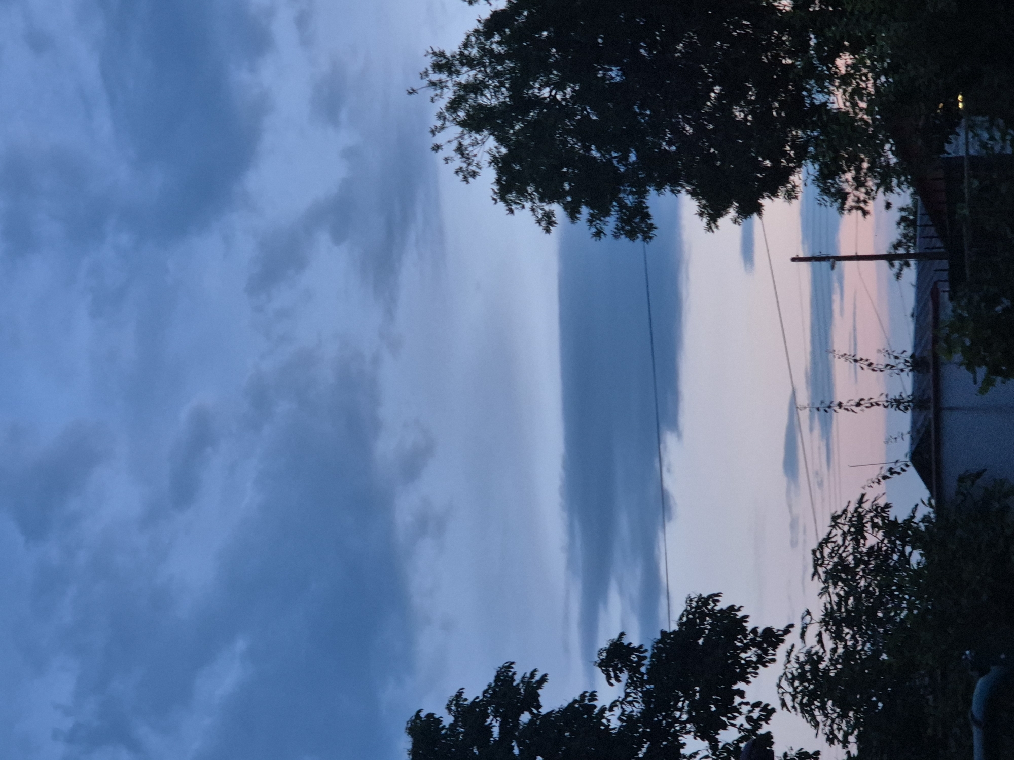 The image shows a twilight sky with scattered clouds over a dark silhouette of trees and rooftops. The sky transitions from a deep blue at the top to lighter shades of pink and blue near the horizon, suggesting either sunrise or sunset. Several power lines are visible crisscrossing the lower portion of the sky. The foreground is dominated by the dark foliage of trees and the dark shapes of buildings. No people or specific actions are depicted. The location is identified as Unknown, Unknown.