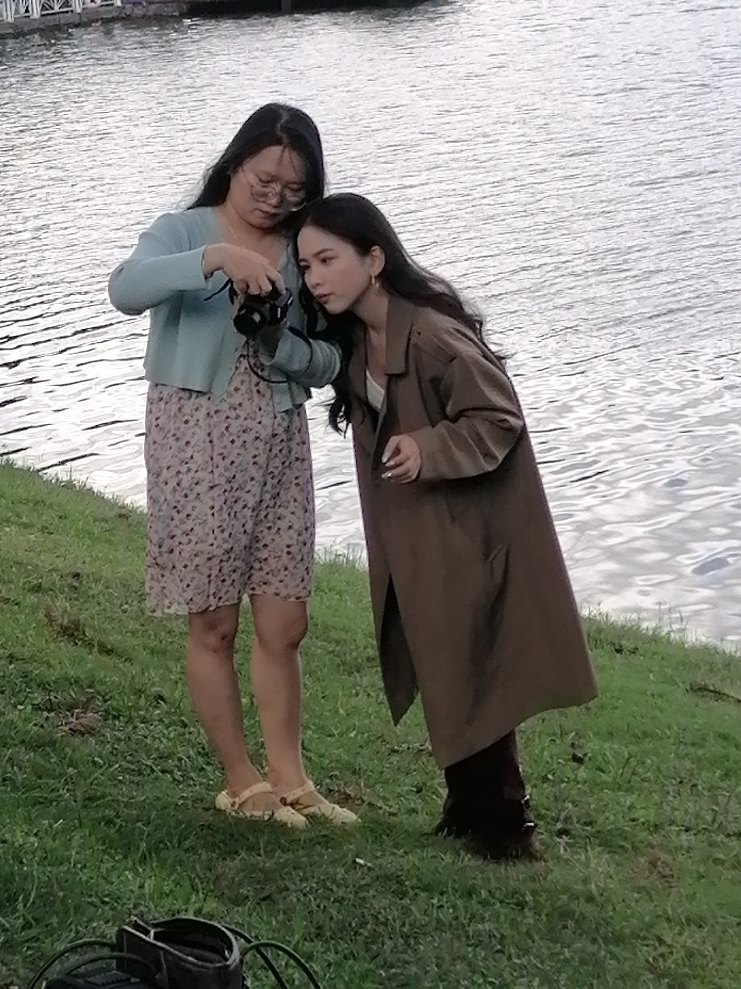 Two young women are standing on a grassy bank next to a body of water, likely a lake or river. The woman on the left, wearing a light blue cardigan over a floral print dress and beige clogs, is holding a black camera and appears to be taking a picture of the woman on the right. The woman on the right is wearing a long, brown trench coat over dark pants and dark boots. Her head is tilted down as she looks towards the camera. The background shows rippling water with a distant structure that appears to be a bridge or pier. The scene is set outdoors with lush green grass in the foreground. The presence of a lake and hilly terrain suggests a location such as Xuan Huong Lake in Da Lat, Vietnam, which is known for its scenic beauty and walking paths.