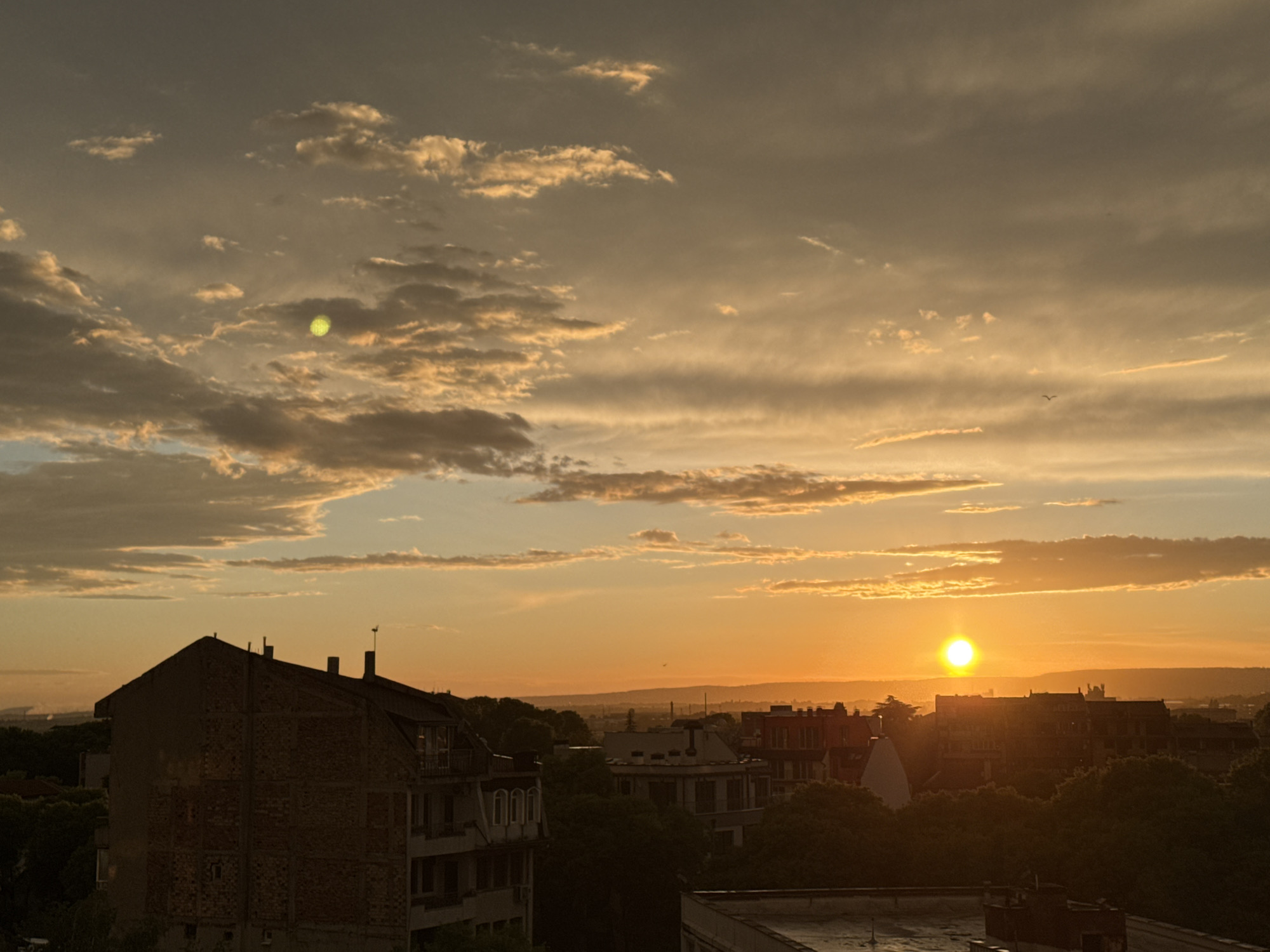 The image shows a sunset over Varna, Bulgaria. The sun is near the horizon, appearing as a bright, yellow-orange disc. Clouds are scattered across the sky, some tinged with orange and yellow hues. Below the horizon, the silhouette of a cityscape with buildings and trees is visible. A large building is on the left side of the image. There is a bright greenish spot in the upper-left of the frame. A bird is flying in the upper-right section of the sky.