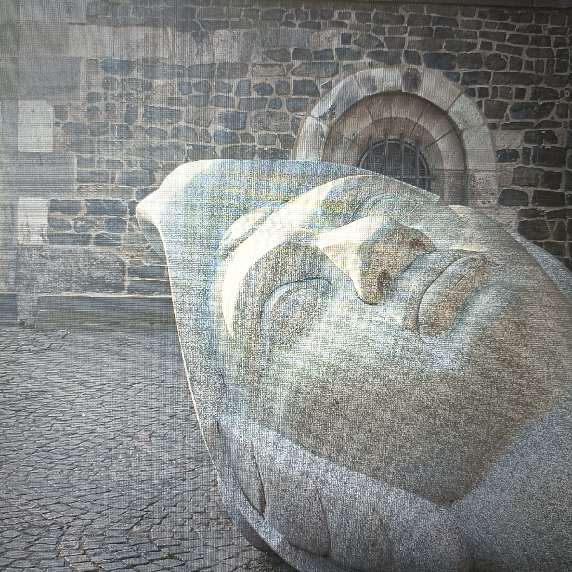 The image depicts a close-up, low-angle view of a large, stylized stone sculpture of a human head, resting on a cobblestone plaza. The sculpture is light gray and appears weathered, with smooth, curved lines defining its features, including closed eyes, a prominent nose, and lips. Sunlight illuminates the left side of the sculpture, casting shadows on the right.
In the background, a textured stone wall of a building is visible, constructed from irregular, dark gray stones. A small, arched window with a metal grate is set into the wall above and to the right of the sculpture. The ground beneath the sculpture is paved with gray cobblestones arranged in a repeating pattern.
The sculpture is located in the city of Rüsselsheim am Main, Germany, specifically in front of the former Adam Opel AG main gate, which is now a part of the Opelvillen.