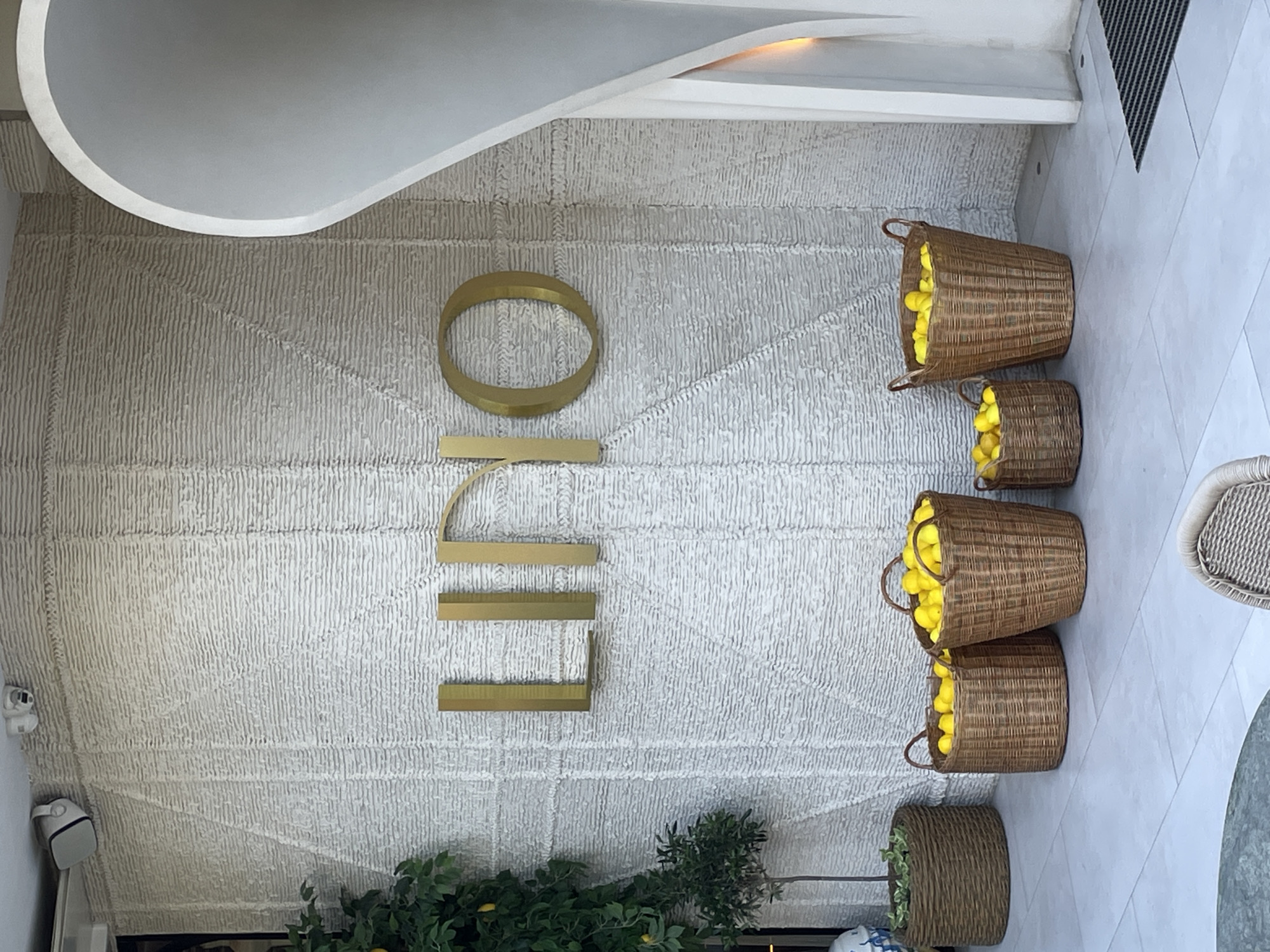 The image depicts the facade of a establishment, likely a cafe or restaurant, with a textured white wall featuring the gold-colored lettering "LINO" prominently displayed. In front of the wall, several woven baskets are filled with bright yellow lemons. To the left, a small tree with green leaves and a few yellow fruits is potted in a woven planter. Two white security cameras are visible mounted on the upper part of the wall. To the right, a curved white architectural element forms an entrance or alcove. The ground is paved with light-colored tiles, and a black doormat is positioned near the entrance. A glimpse of a metal chair and table is visible in the lower foreground. The scene suggests an outdoor seating area. The location is identified as Sochi, Russia.