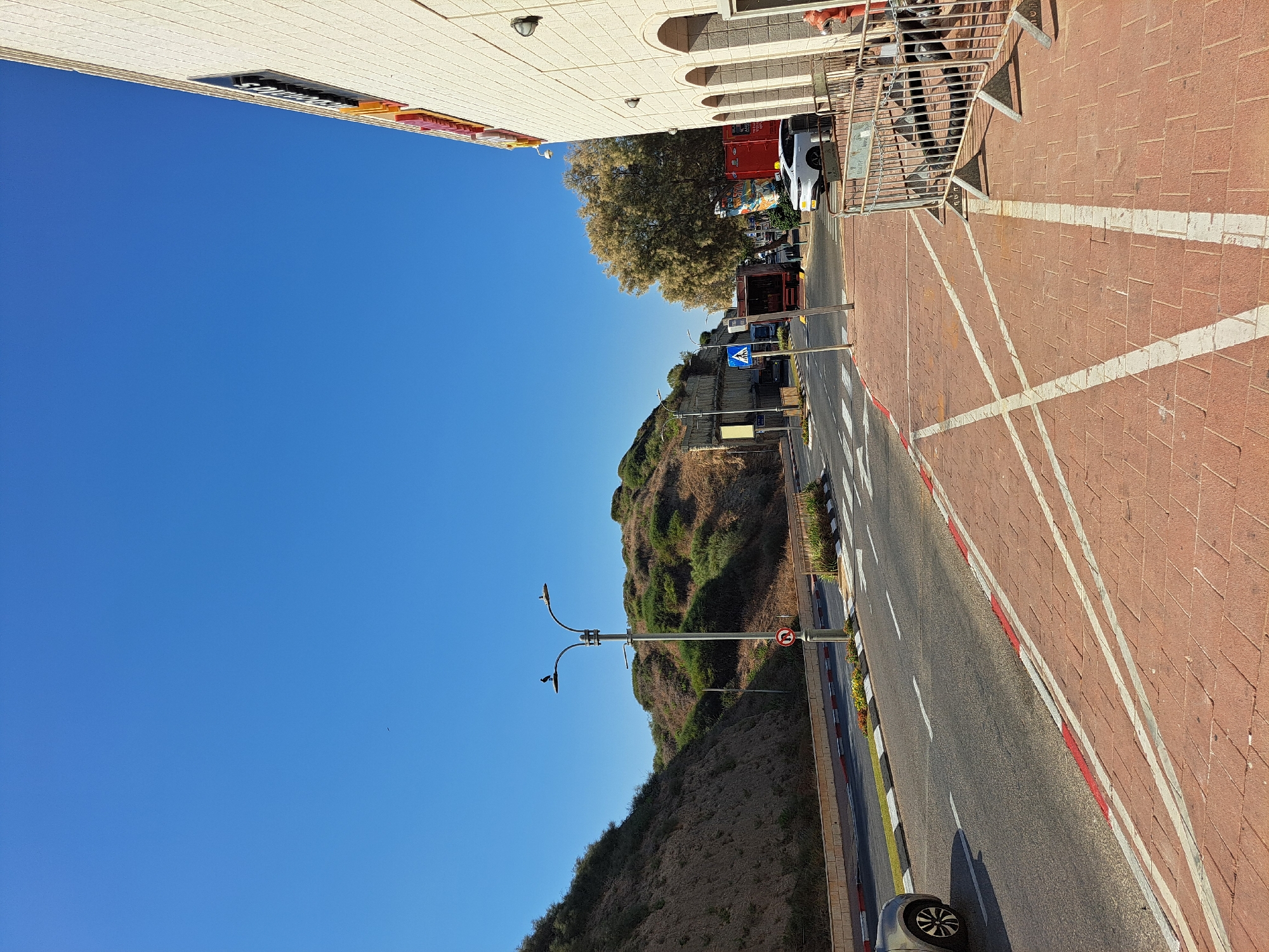 The image captures a street scene in Herzliya, Israel, on a clear, sunny day. The foreground is dominated by a red brick sidewalk with white painted lines indicating parking spaces. A metal barrier is erected along the edge of the sidewalk, separating it from the street. A red fire hydrant is visible near the barrier. The street itself is paved with asphalt, marked with white lane lines and a red and white curb. A silver car is seen driving on the left side of the road.
In the middle ground, a gentle slope covered with dry vegetation forms a hillside on the left. A street lamp with a bird perched on it stands near the road. A road sign with a red circle and diagonal bar indicates a prohibition. Further down the street, a pedestrian crossing is marked on the asphalt. Traffic signals and utility poles are also visible. Several vehicles, including a red truck and a white car, are parked or driving on the street. Trees with light-colored foliage are present on the right side of the street.
The background features more of the vegetated hillside under a bright blue sky. On the far right, the side of a building with light-colored stone cladding and arched windows is visible. A sign with lettering is mounted on the building. The overall impression is of a sunny day in a well-developed urban environment with elements of natural landscape.