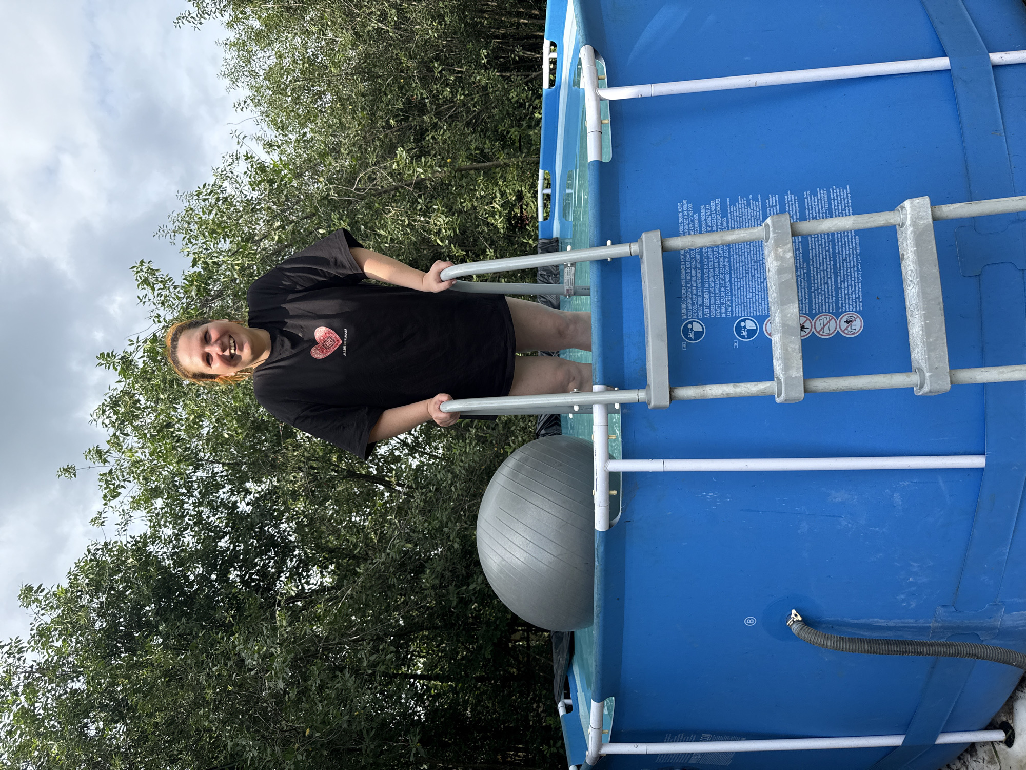 An overhead shot captures a woman with light brown hair, wearing a black t-shirt with a red heart graphic and text that reads "MADE IN RUSSIA", standing in a blue, above-ground swimming pool. She is smiling and looking at the camera. Her legs are submerged in the pool water. A grey exercise ball is partially submerged in the water next to her. A silver ladder with grey rungs extends into the pool, and the woman's right hand is resting on one of the ladder's rails. The pool is surrounded by green trees and vegetation under a cloudy sky. The scene appears to be in a backyard in the village of Khokhryaki, Russia.