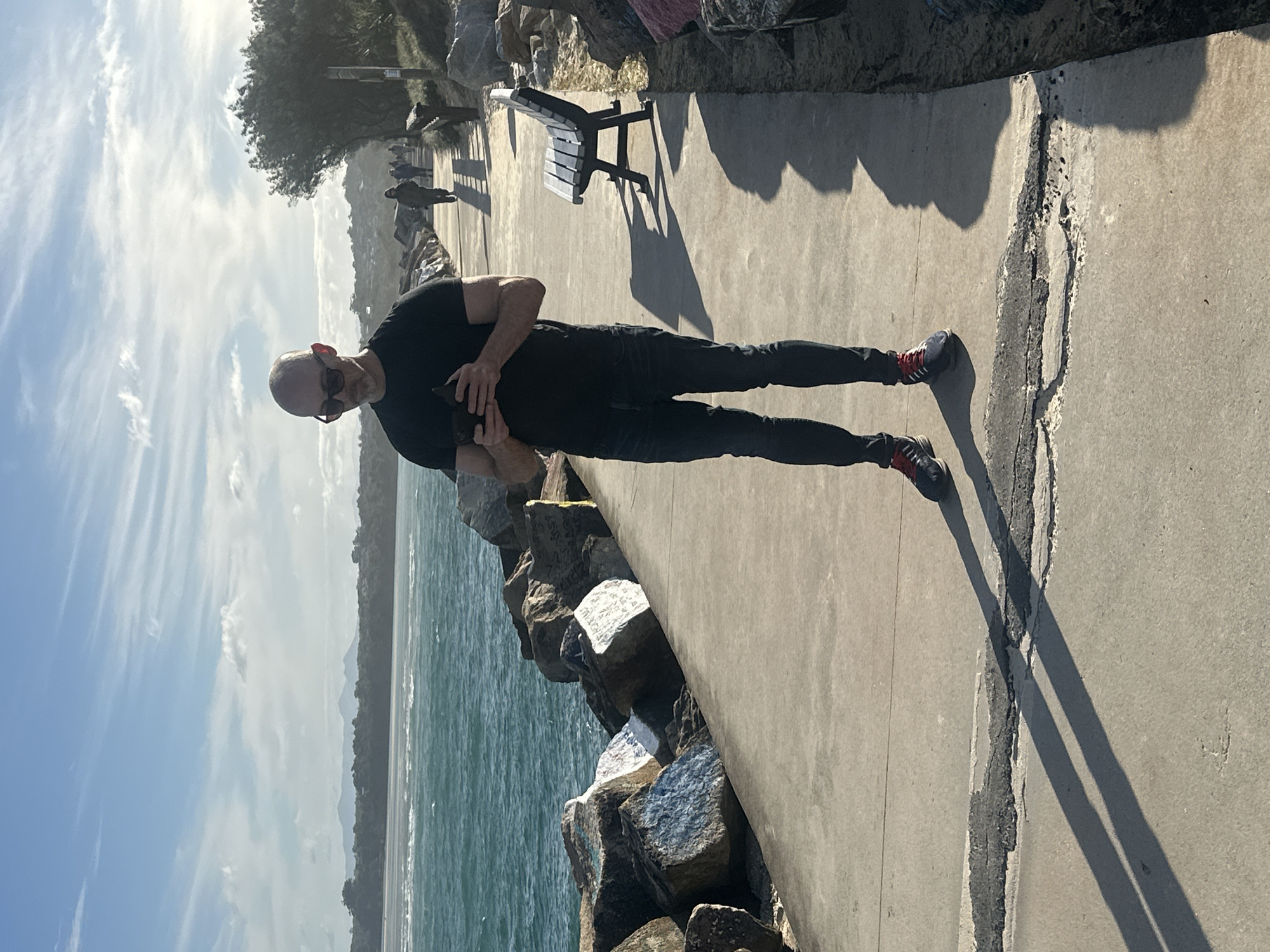 The image depicts a man standing on a concrete walkway next to a rocky shoreline in Nambucca Heads, Australia. The man, bald and wearing sunglasses, is looking down at a black smartphone he holds in his hands. He is dressed in a black t-shirt and dark blue jeans, with athletic shoes. The walkway is lined with large rocks, some of which have graffiti on them, leading towards the ocean. The sea is a vibrant turquoise, with waves visible. In the background, other people are walking along the path, and a park bench is situated on the right side of the walkway. The sky is blue with scattered white clouds, and the sun is casting long shadows on the ground. The landscape in the distance suggests a coastal town with hills and vegetation.