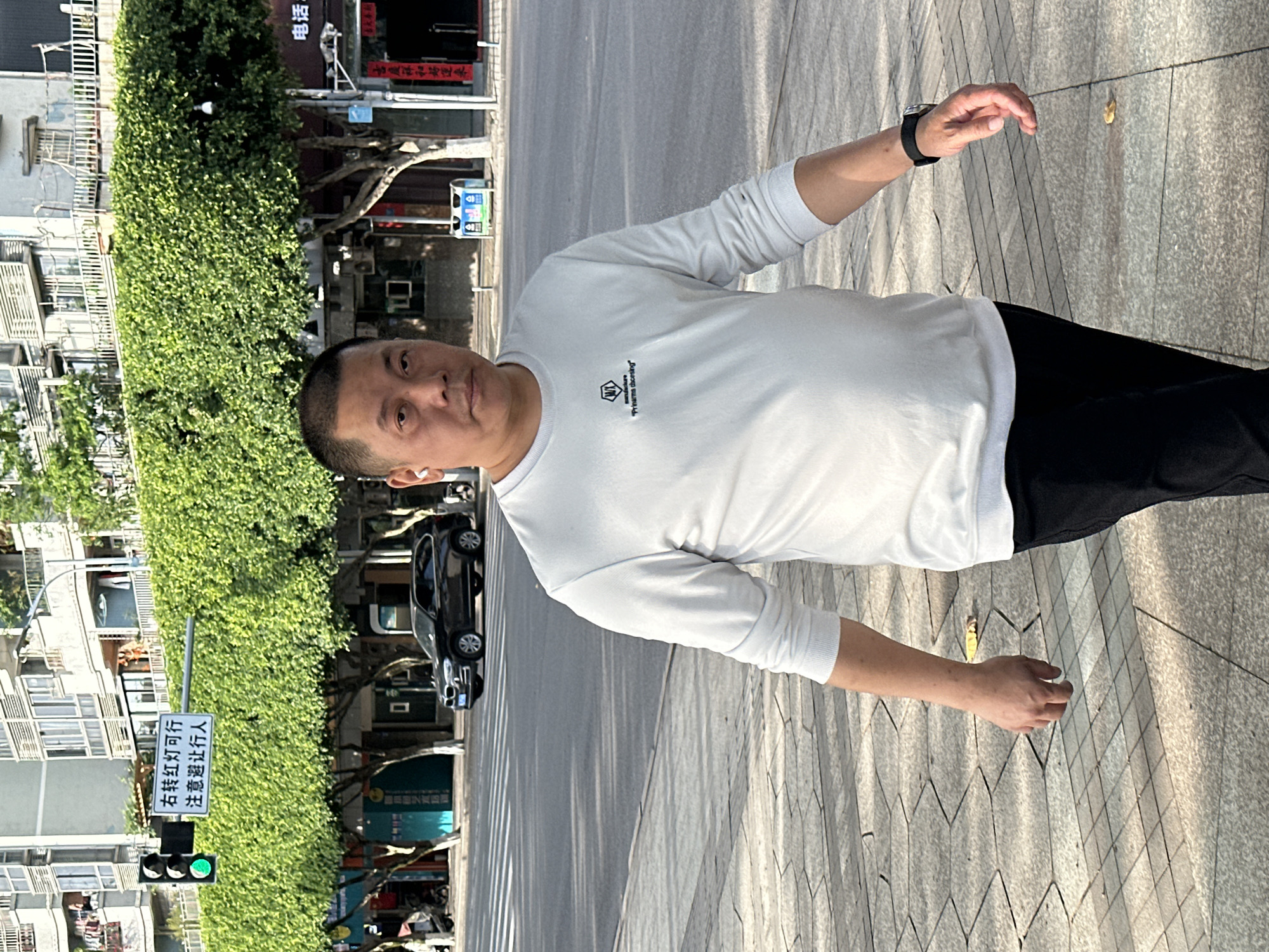 A man is captured standing on a paved sidewalk in an urban environment during clear, sunny daylight hours. He is looking directly at the camera with a neutral expression. The subject wears a white long-sleeve t-shirt with a black logo on the chest, featuring a stylized 'W' within a shield shape, and the text "YI WANG Recording" beneath it. He is also dressed in dark pants and has a black wristwatch on his left wrist, with an earbud visible in his left ear.
Behind the man, a street stretches parallel to the sidewalk, with a black car partially visible further down the road. Green bushes and foliage line the street, behind which multi-story buildings with windows are discernable. A traffic light showing a green signal and a street sign with Chinese characters are prominent on the left side of the frame. The sign reads "右转红灯可行 注意避让行人," which translates to "Right turn on red light is permitted, please give way to pedestrians." The sidewalk features patterned paving stones, and a single yellow leaf is visible on the ground, suggesting a possible seasonal detail. The scene depicts an outdoor urban setting, consistent with Jiangjin, China.