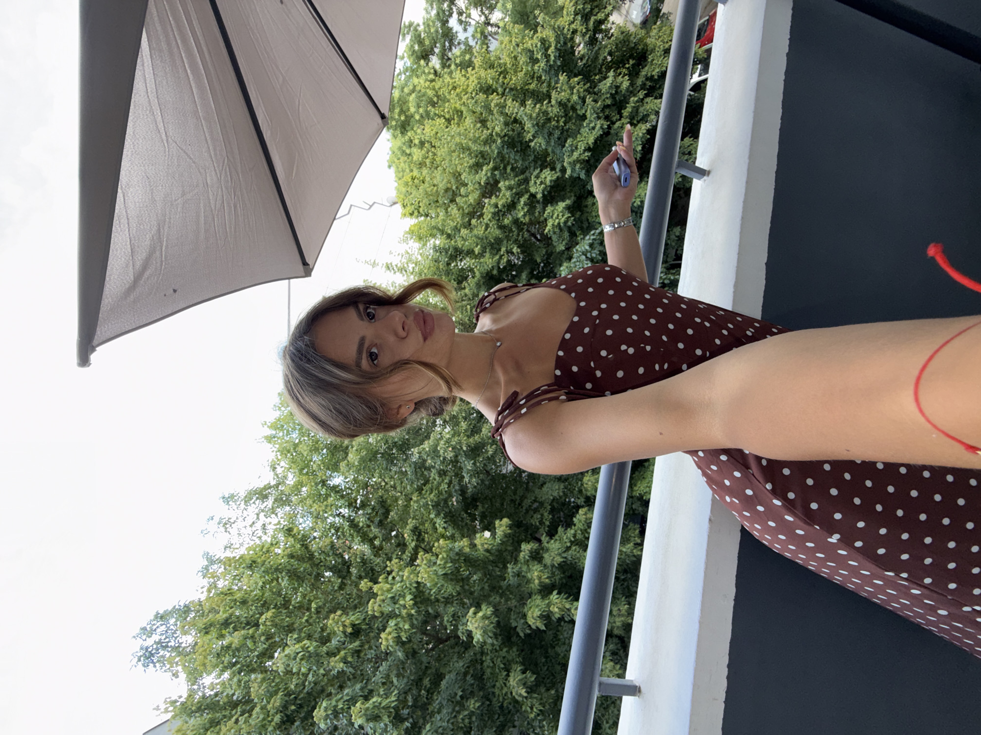 A young woman stands on a balcony, holding a phone in her right hand. She is wearing a brown polka-dot dress. Behind her, lush green trees fill the background, suggesting an urban setting, possibly Plovdiv, Bulgaria, given the context. A large, light-colored umbrella is partially visible in the upper right corner of the frame. The woman is looking towards the camera with a slight pout. The balcony railing is visible in the foreground, with a white ledge above a dark gray wall. The sky is overcast.