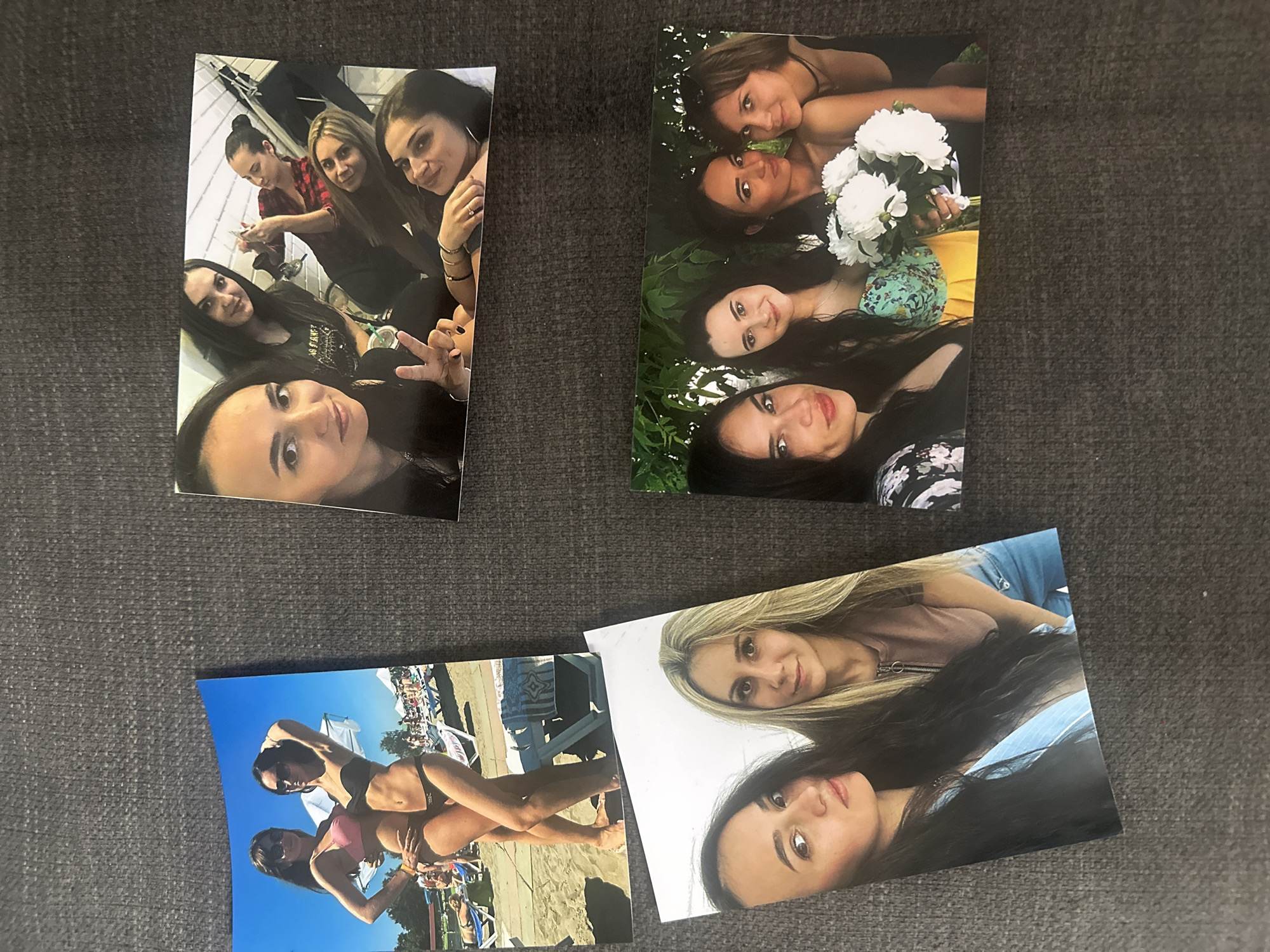 Four photographs are laid out on a dark gray, textured surface. The photographs depict groups of young women, likely friends, in various casual settings.
The top left photograph shows four women. One in a red plaid shirt is in the background, looking at her phone. To her right, another woman with blonde hair smiles at the camera. Next to her, a woman with dark hair and hoop earrings makes a peace sign with her fingers. In the foreground, a woman with dark hair and bright eyes looks directly at the viewer. The setting appears to be indoors, possibly a bathroom or changing room, with white tiled walls visible.
The top right photograph features five women outdoors. Three are in the foreground, with two of them holding a bouquet of white flowers. They are smiling and posing for the camera. The background shows lush greenery.
The bottom left photograph shows two women in bikinis posing on a beach. They are both wearing sunglasses. The background indicates a sunny day at a beach resort, with lounge chairs and other people visible.
The bottom right photograph shows two women, one with blonde hair and the other with dark hair, looking at the camera. One is wearing a light pink top with a zipper detail, and the other has dark hair. The setting is unclear, but appears to be outdoors.
The photographs suggest a collection of casual memories from outings and gatherings among friends. The overall mood is relaxed and joyful. No visible text is present in any of the photographs.