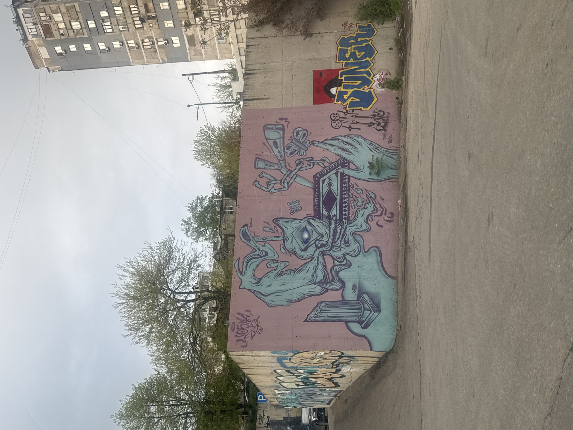 The image displays an urban outdoor scene in Tbilisi, Georgia, centered on a large graffiti mural covering a concrete wall. The wall, painted in a pinkish-mauve hue, stands alongside an asphalt road. In the background, a multi-story building with many windows and air conditioning units is visible under an overcast, bright sky. Several trees with light green foliage or bare branches are scattered throughout the landscape, suggesting spring or early autumn.
The primary subject is the intricate and colorful graffiti mural. It features a surreal, stylized creature or head with a single large eye, rendered in shades of light blue, purple, and white, with flowing, smoke-like elements. A human-like hand, also in light blue, extends from the right side, appearing to hold or interact with a geometric, patterned box-like object. Other elements integrated into the design include butterflies, abstract shapes, and a classical-style column.
Above the main mural, another graffiti piece in bold, blocky blue and yellow letters spells "SUMMER," superimposed on a red rectangular background that contains a small, dark, stylized figure. Below the main artwork, a smaller tag includes the text "TBN" and the year "2020." Further down the wall and around a corner, more graffiti tags are visible.
The scene captures a static moment during daylight hours. There is no specific activity taking place, but the vibrant street art creates a dynamic visual element within the concrete urban environment.