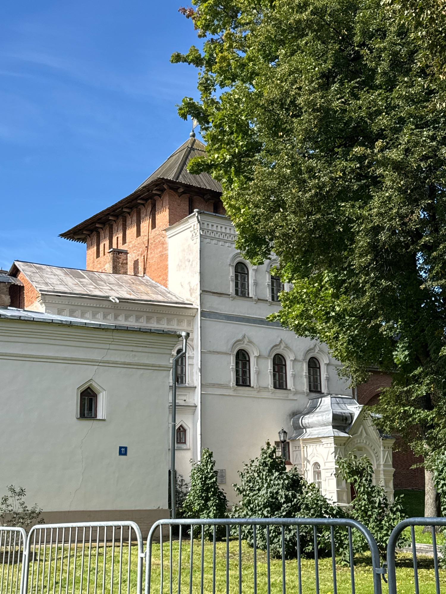 The image shows a bright, sunny day in Veliky Novgorod, Russia, focusing on a historical building complex. The architecture is a mix of styles, with a prominent red brick tower topped with a wooden roof and decorative corbels. Adjacent to it is a white building with arched windows and decorative stucco work. A smaller white structure with an ornate, multi-tiered roof is also visible. The foreground features a metal fence and manicured green grass, with lush green trees providing shade. The blue sky is clear, indicating good weather. The scene appears to be a public space, possibly a museum or a historical site. A blue house number "11" is affixed to the white wall of the building.