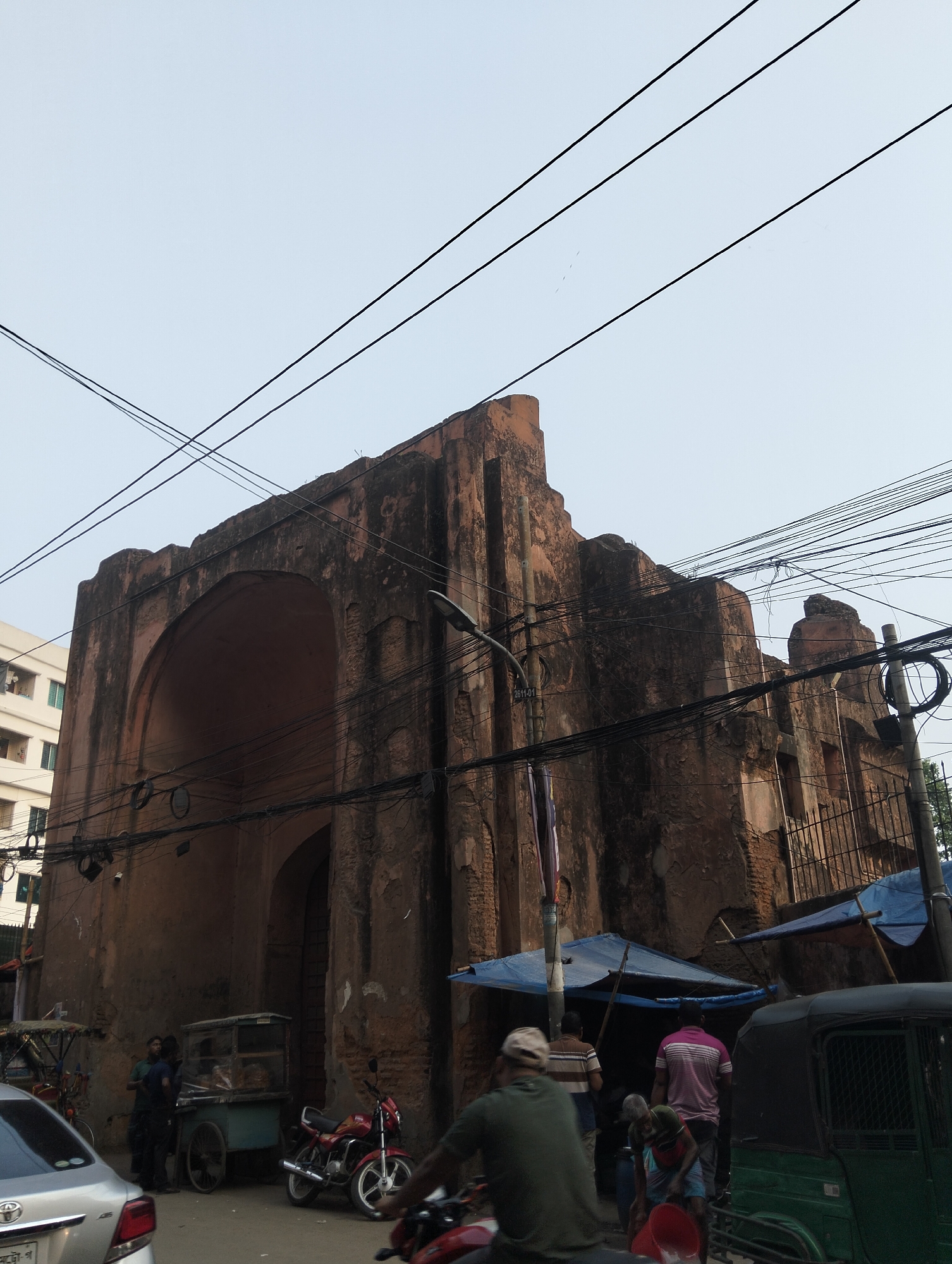 The image depicts the Dhaka Gate, also known as the Mir Jumla's Gate, a historical monument in Dhaka, Bangladesh. The gate, made of brick and stone, is in a state of disrepair, with visible weathering and discoloration. A large arched opening dominates the structure, and electrical wires crisscross the sky above it. Below the gate, there's a busy street scene with various forms of transportation including cars, motorcycles, and auto-rickshaws. Several people are present, including vendors with carts and pedestrians. Some blue tarps are erected, forming makeshift stalls or covers along the side of the gate. The overall environment suggests a congested urban setting.